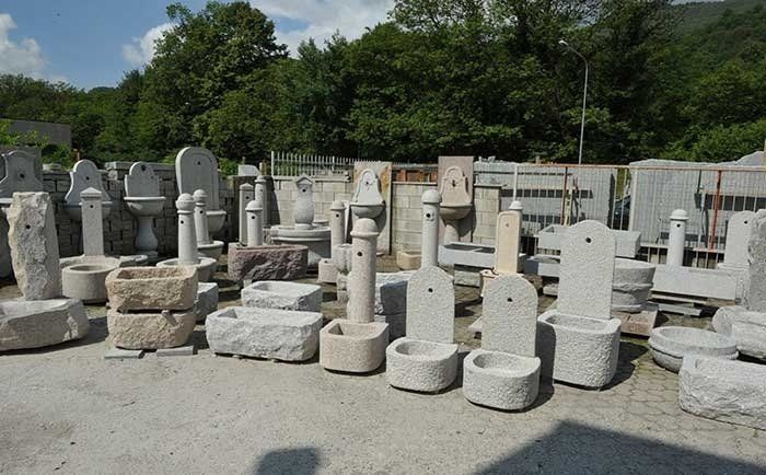 Una selezione di fontane e vasche in pietra scolpita, esposte in un cortile esterno in pietra con alberi sullo sfondo.