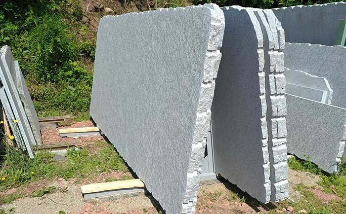 Grandi lastre rettangolari di granito grigio chiaro, posizionate verticalmente all'aperto su supporti di legno in un cortile di pietra.
