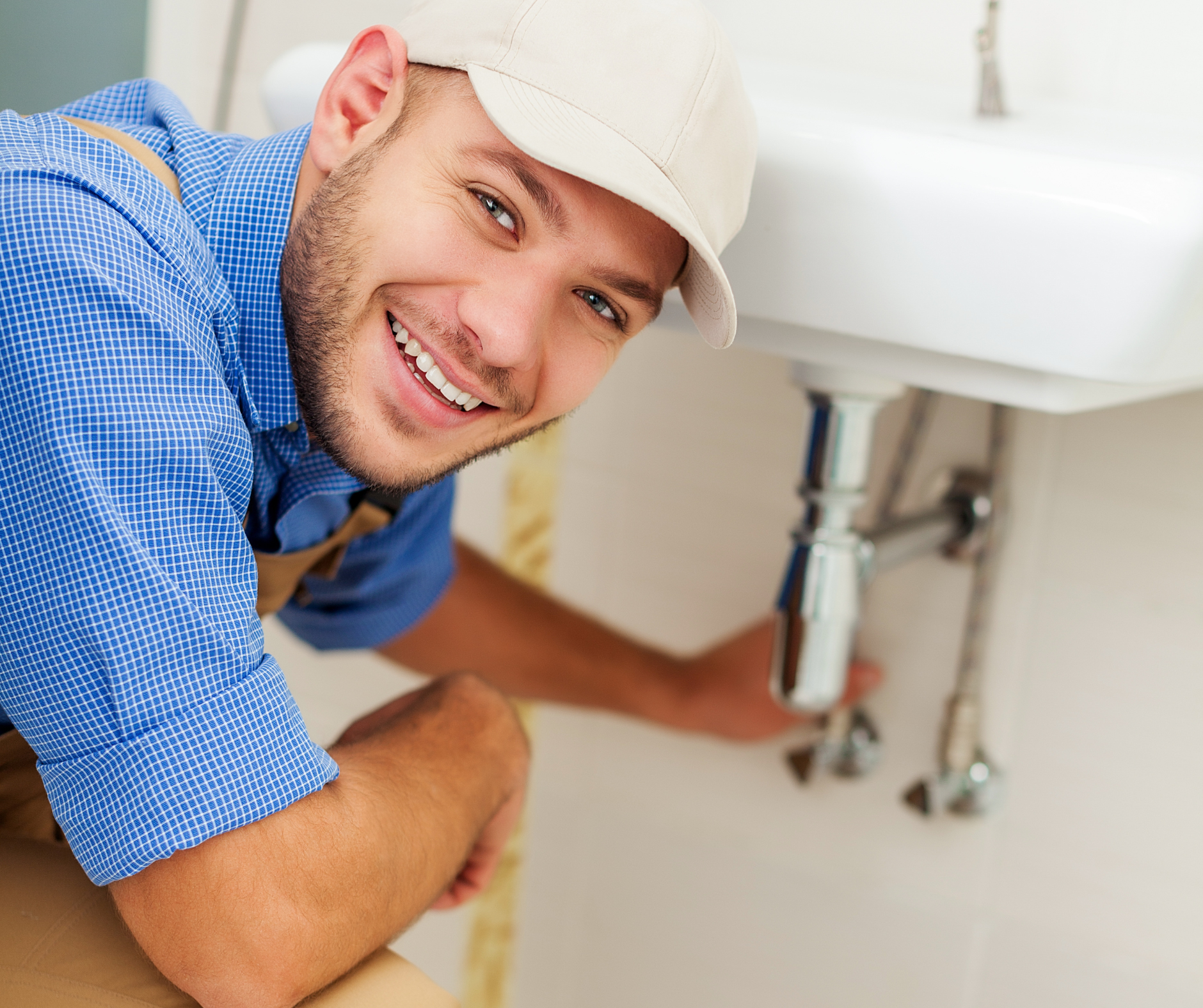 Plumber smiling while working on a sink in a bathroom, wearing a cap and overalls.