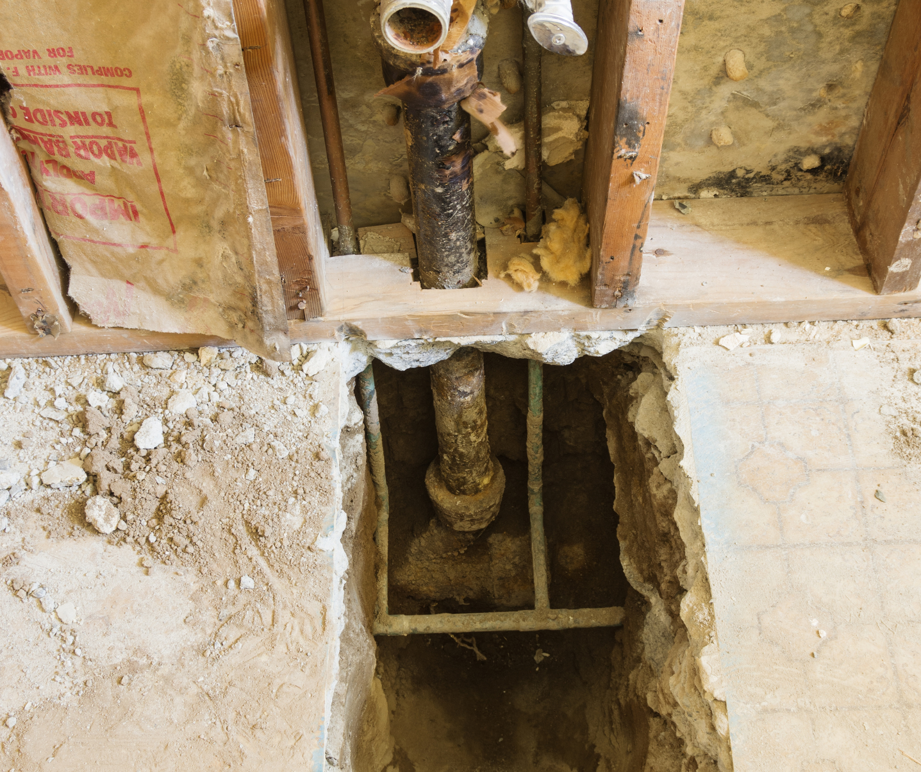 Hole in floor reveals plumbing pipes. Wooden framing and damaged wall visible.