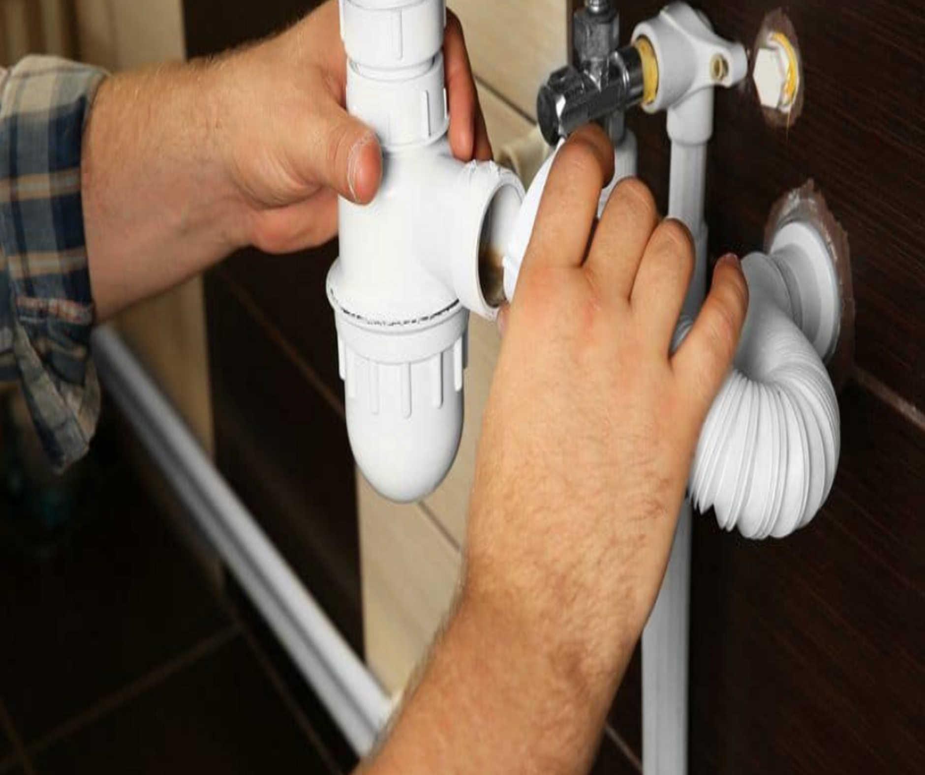 Hands assembling white plumbing pipes under a sink in a bathroom setting.