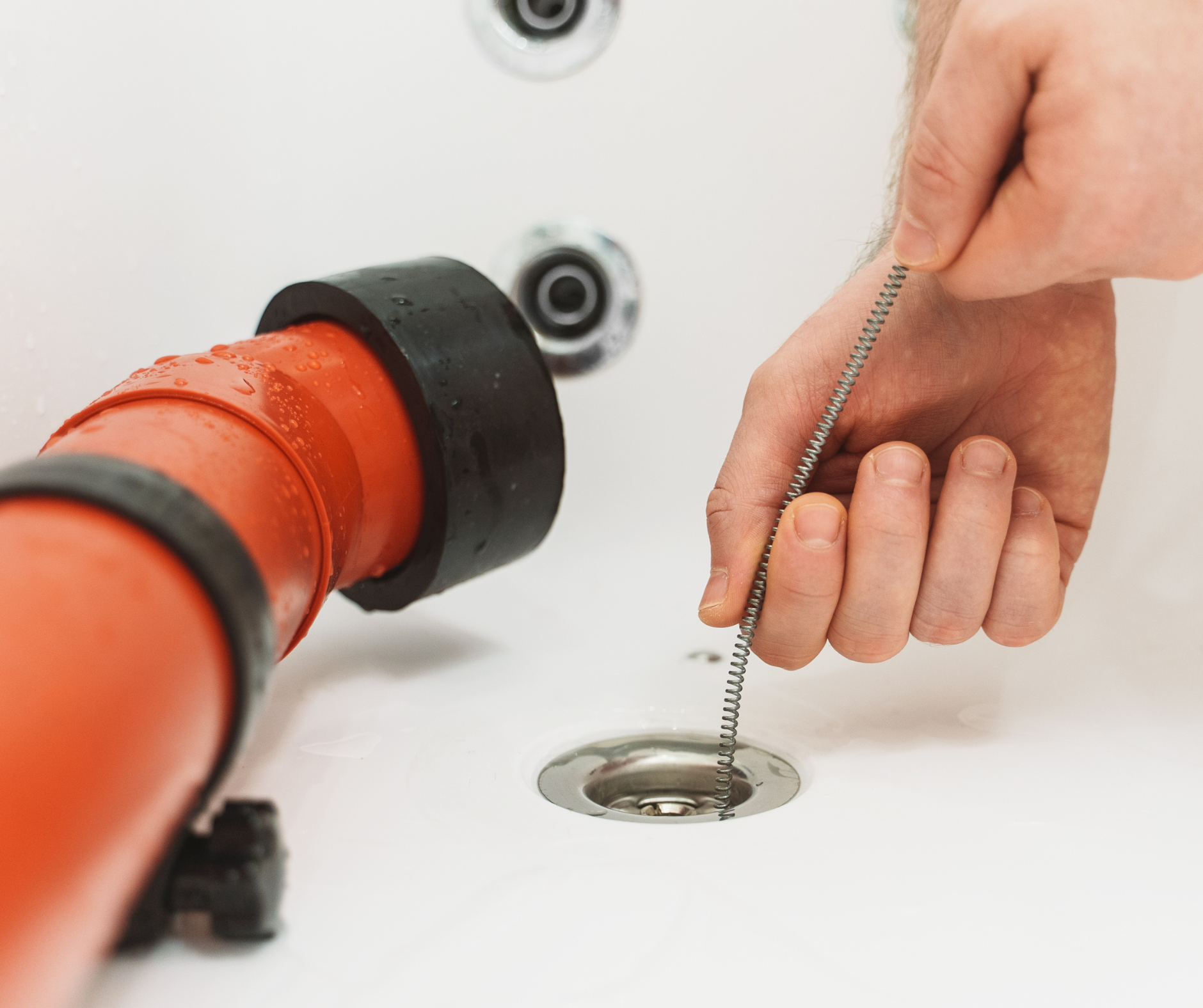 Person using drain snake to unclog a bathtub drain. Close-up with orange and black tools in white bathroom.