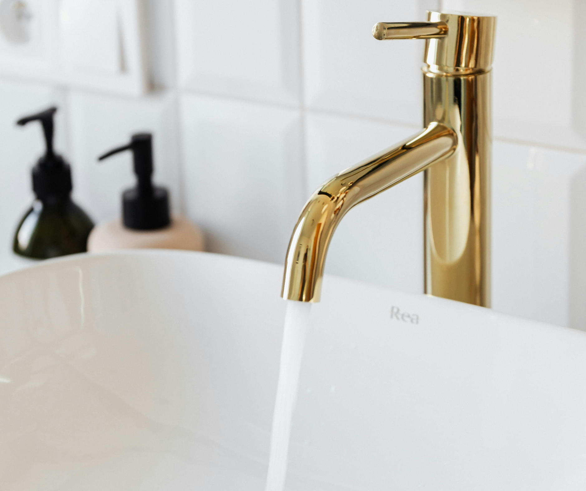 Gold faucet pouring water into a white sink, with soap dispensers in background.
