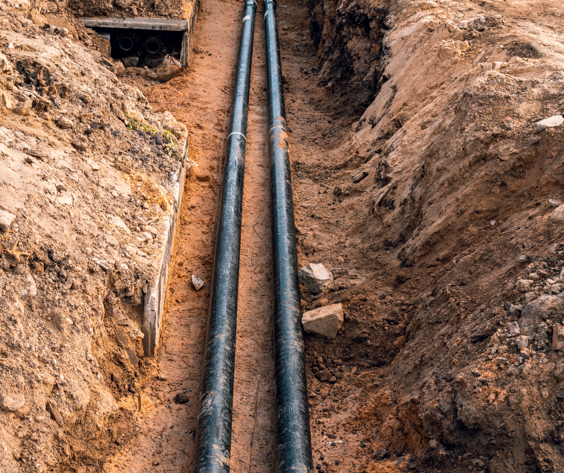 Two black pipes laid in a trench dug in brown soil, ready for burial.