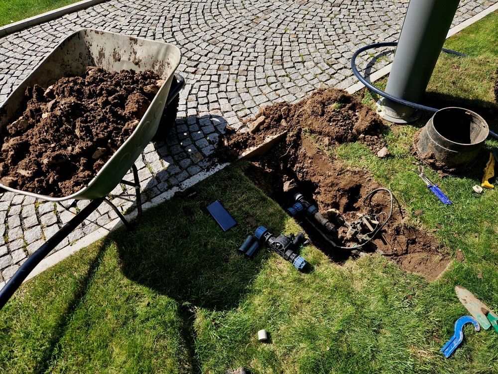 Gardener in green uniform trimming a bonsai tree in a garden setting with various colorful plants.