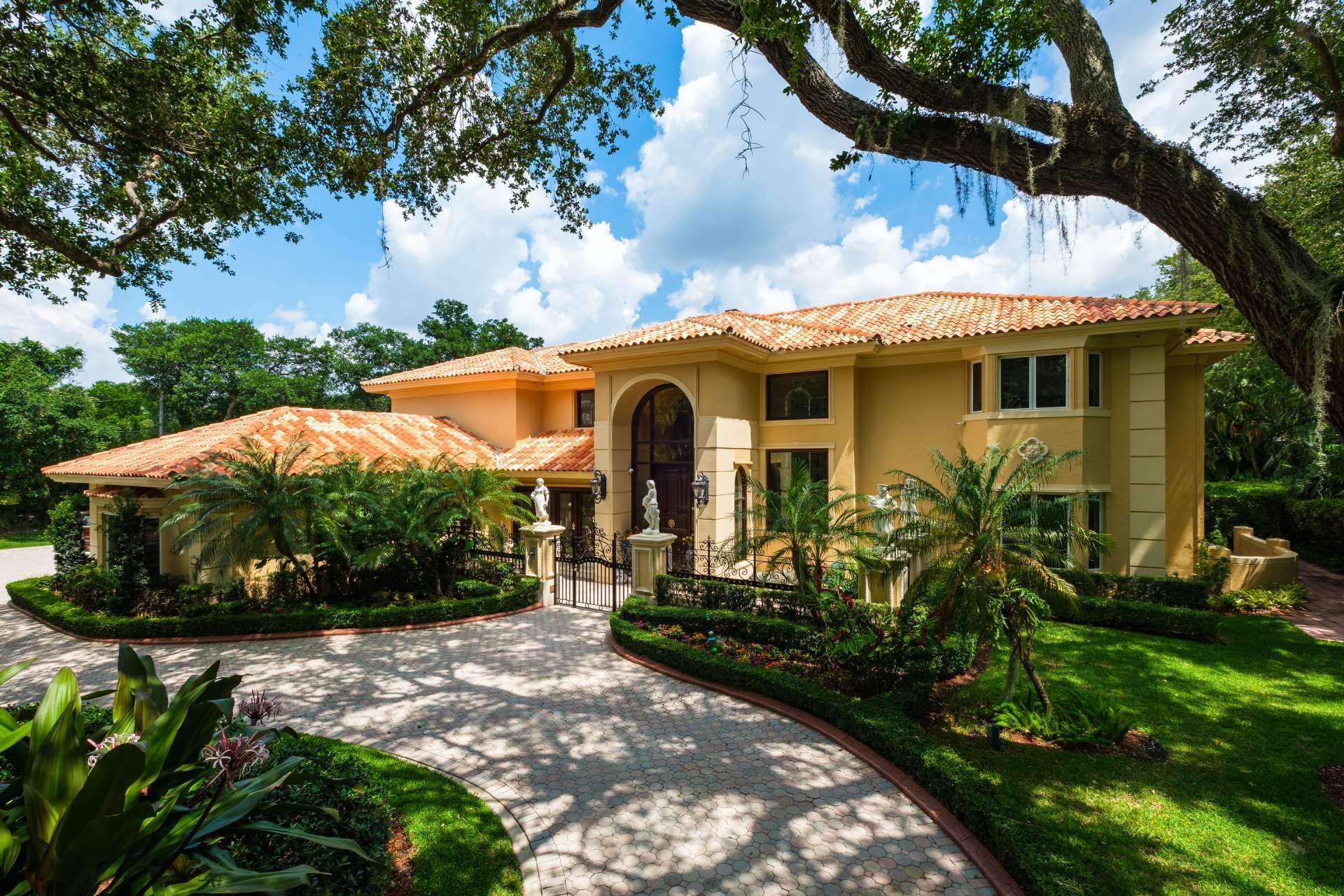 Yellow stucco mansion with red tile roof and circular driveway lined with lush landscaping under a blue sky.