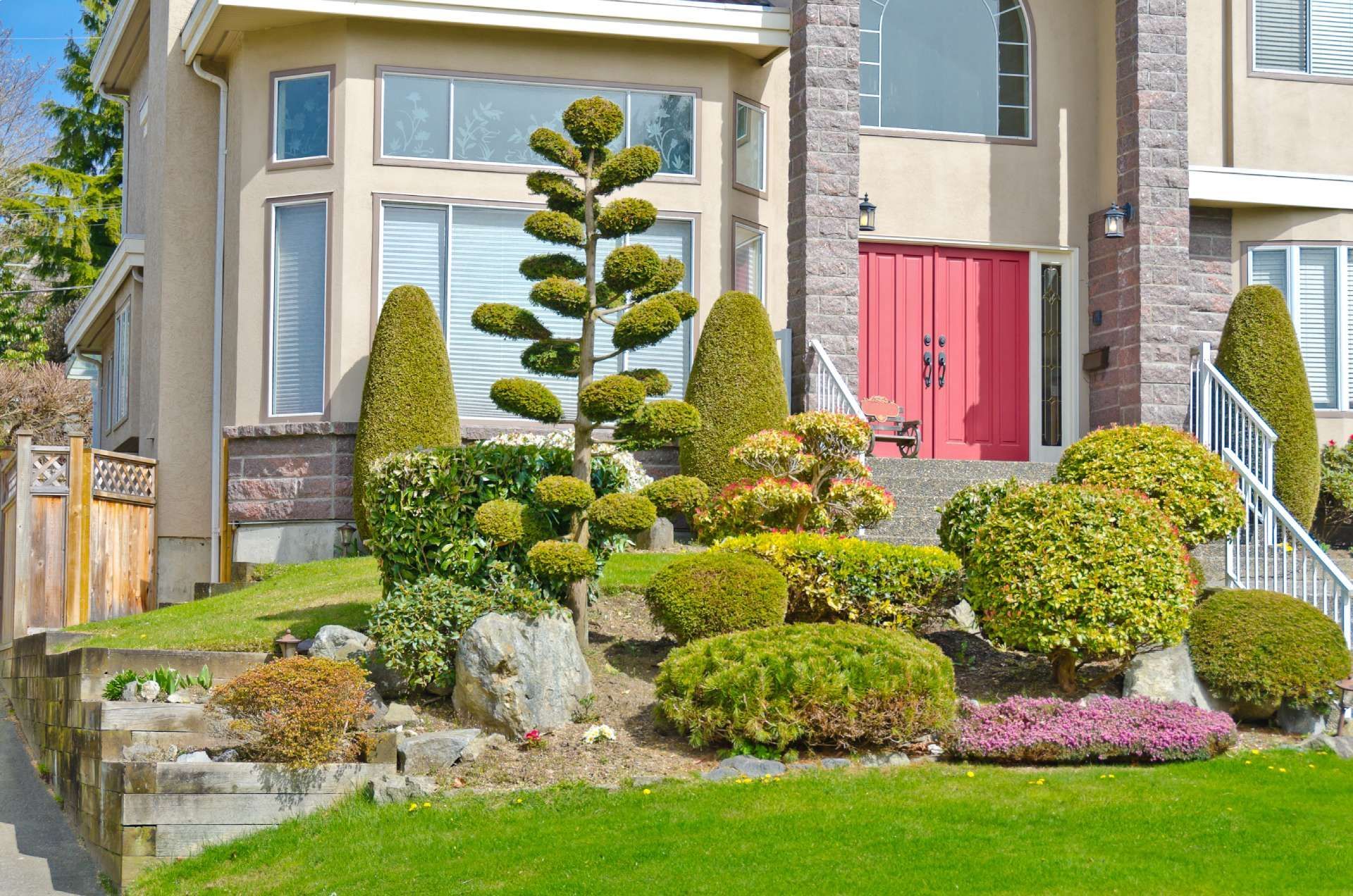 Two-story beige house with red front door, terraced landscaping, topiary, and green lawn.