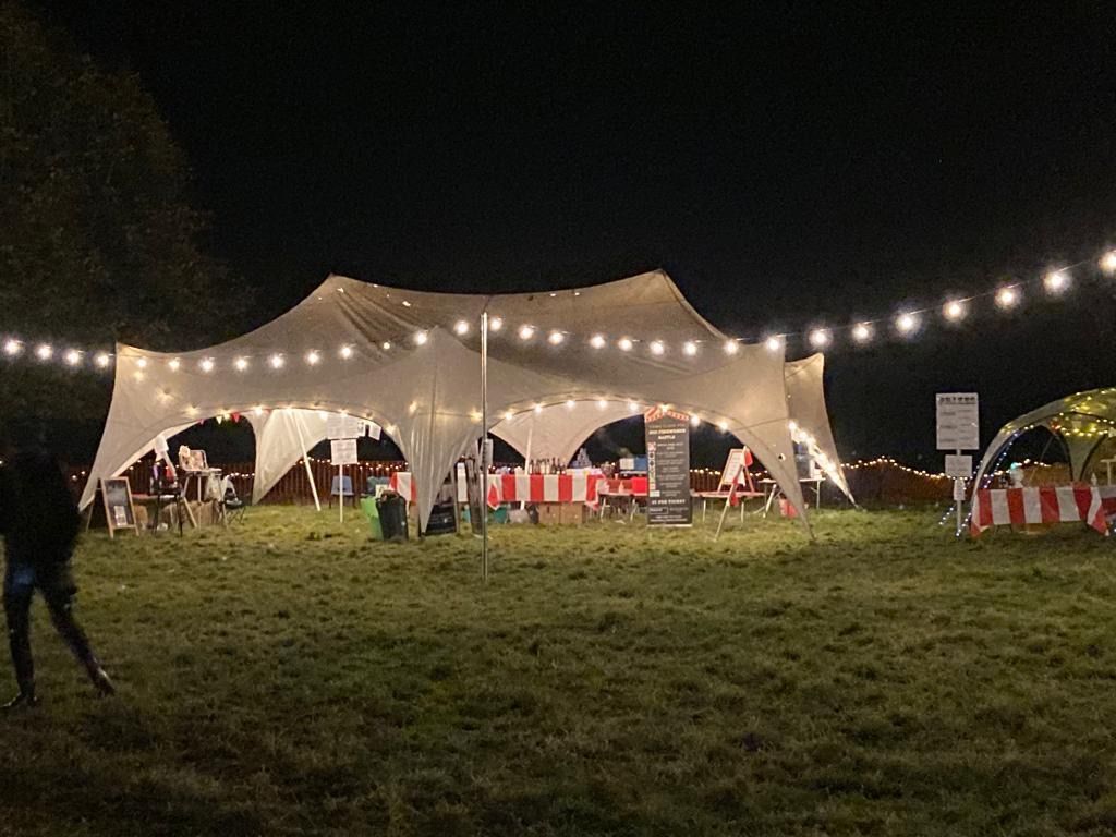 A nighttime outdoor event. Large white tent lit by string lights. People are present.
