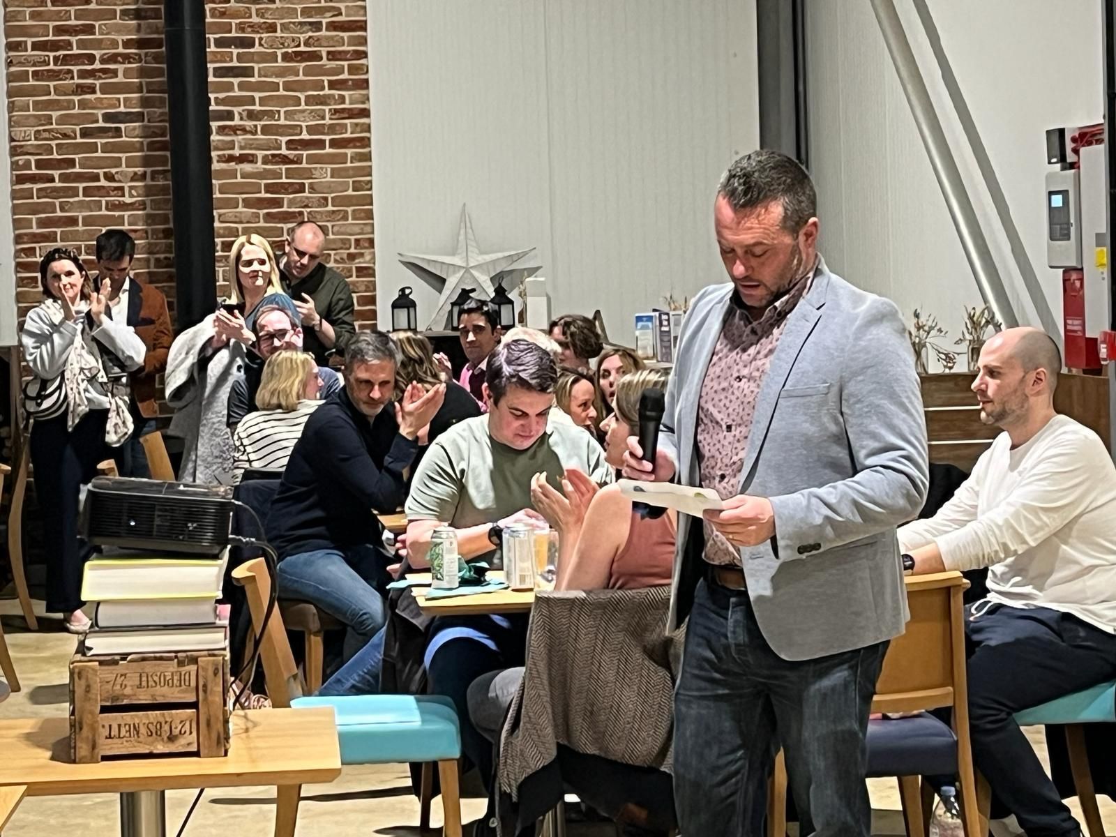 Man speaking at an event, reading from paper. Audience clapping and watching. Interior with tables and brick wall.