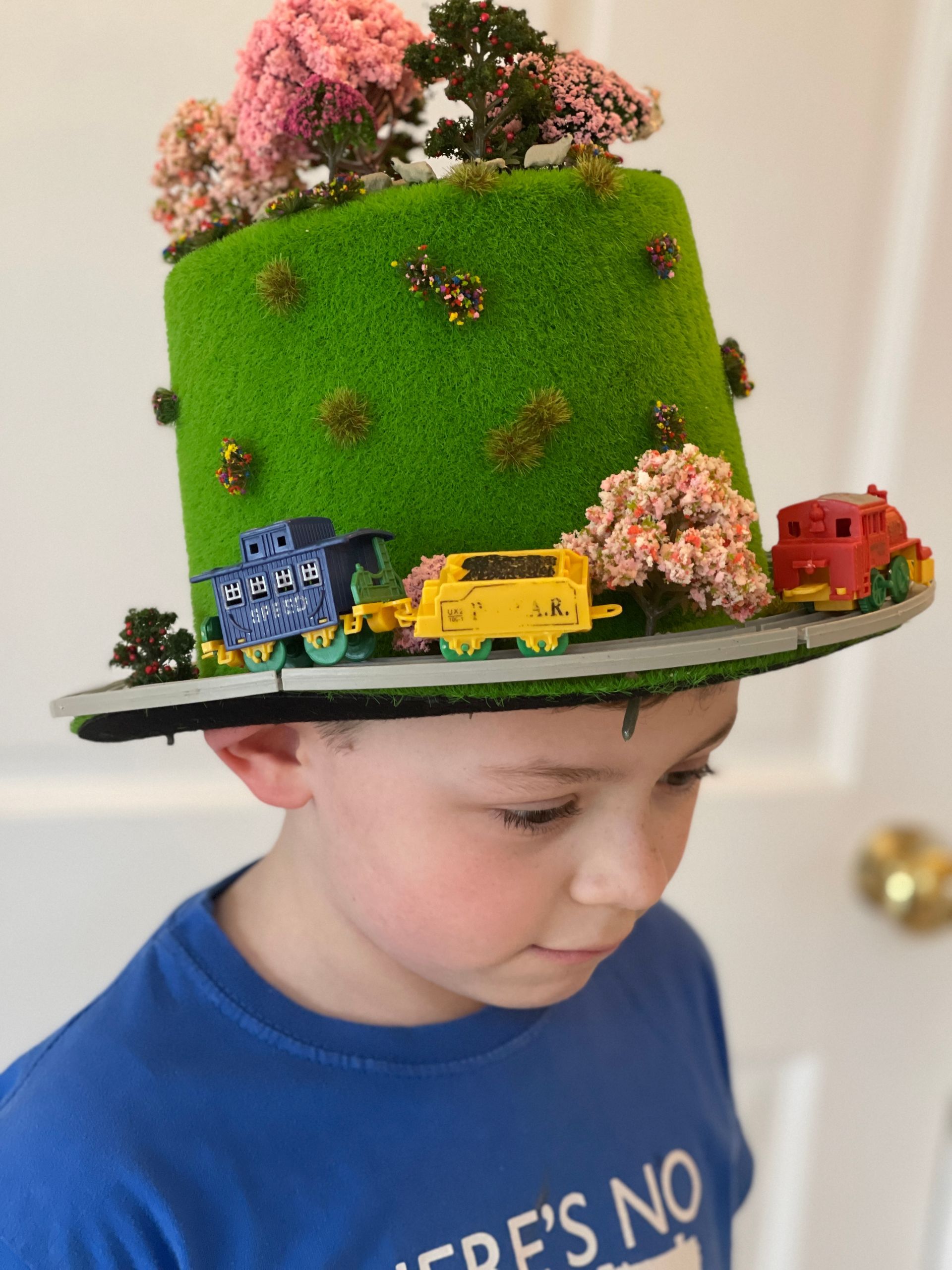 Boy wearing a whimsical green hat decorated with toy trains, trees, and faux foliage.
