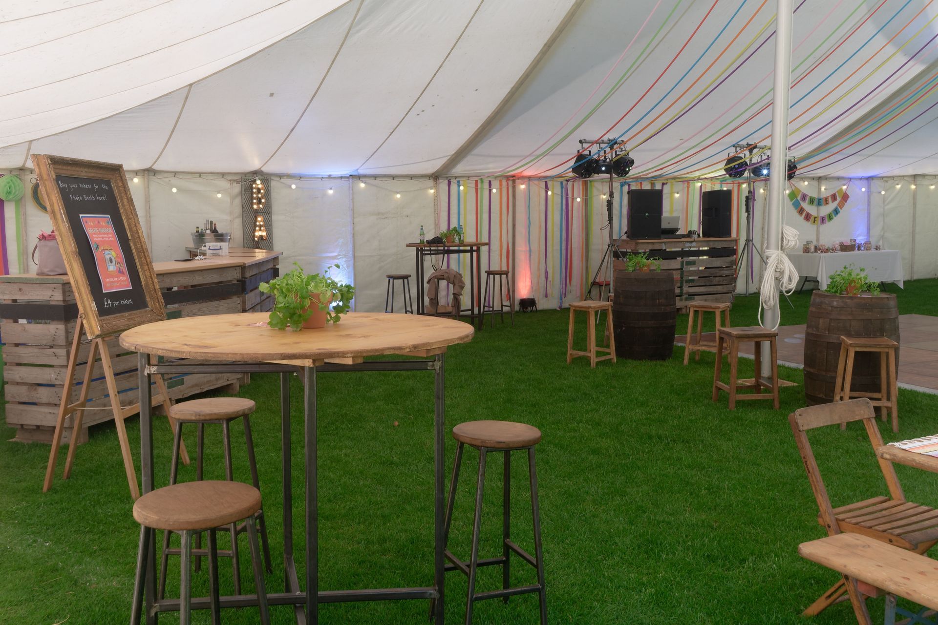 Inside a party tent with wooden tables, stools, and a bar on a grass floor.