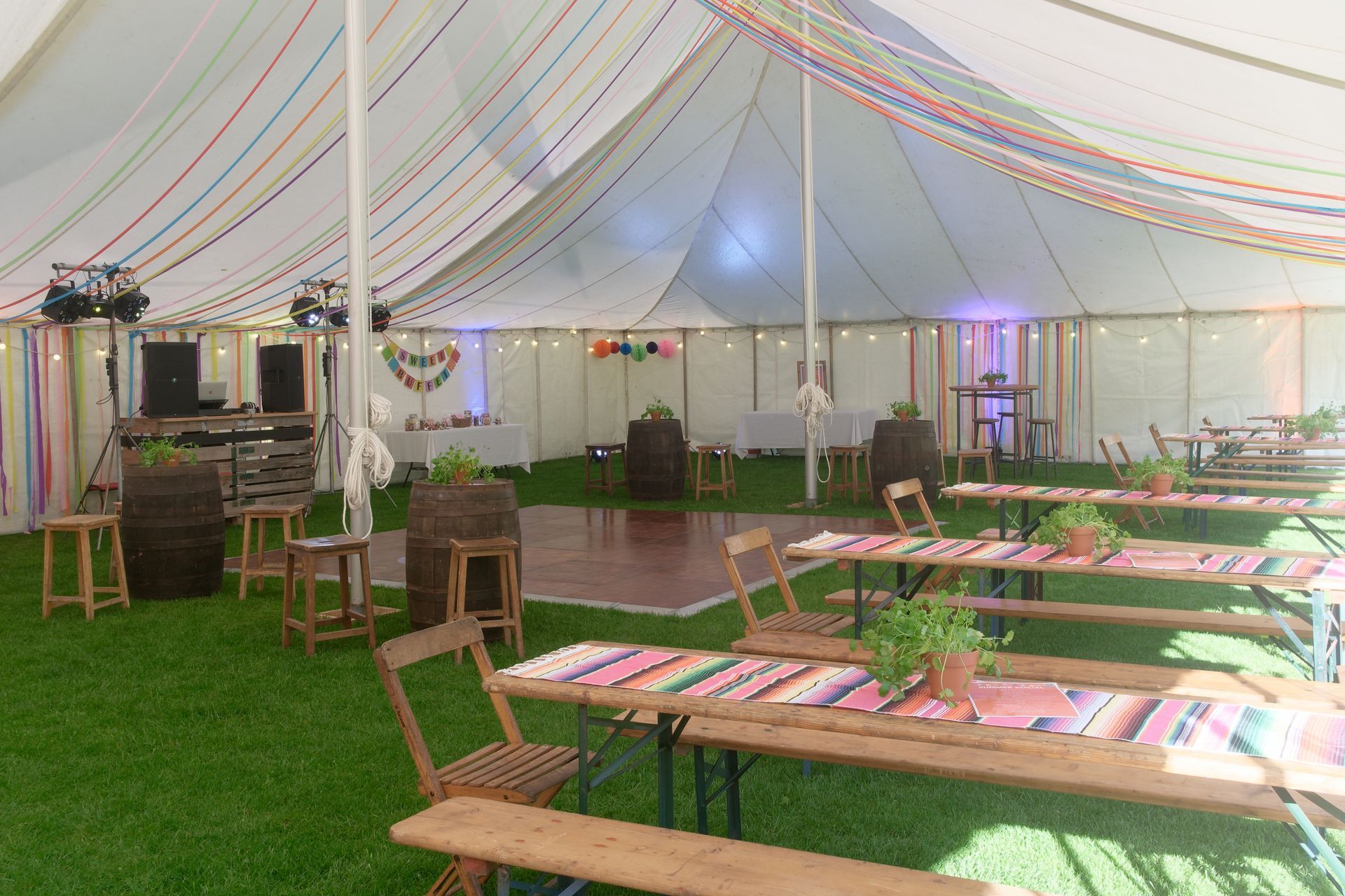 A tent interior with tables, a dance floor, a DJ setup, and colorful decorations; a party or event space.