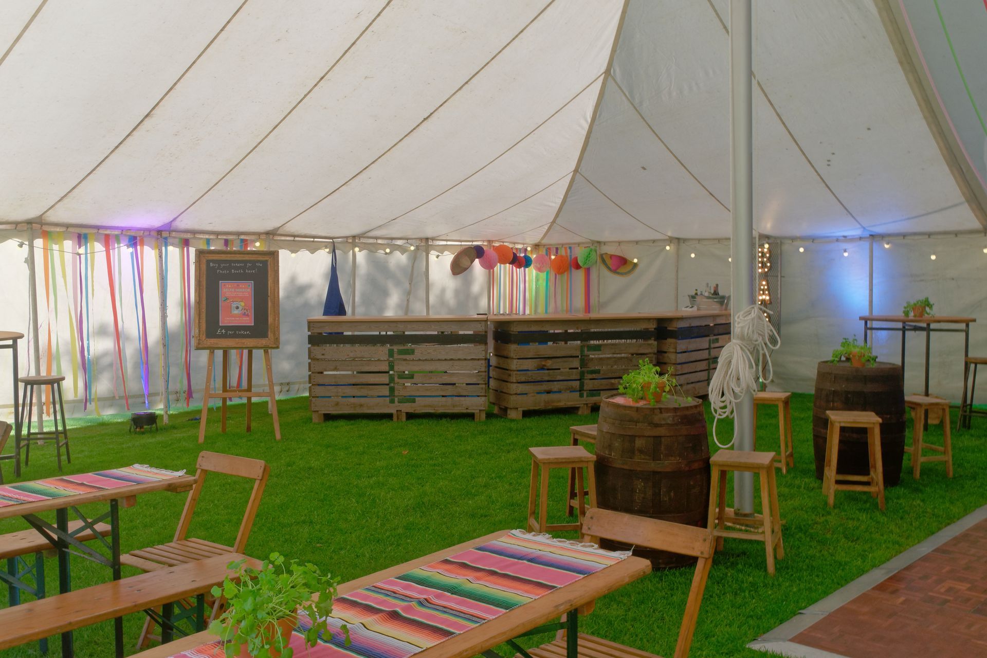 Inside a tent with tables, bar, barrels, and stools on artificial grass, decorated for a party.