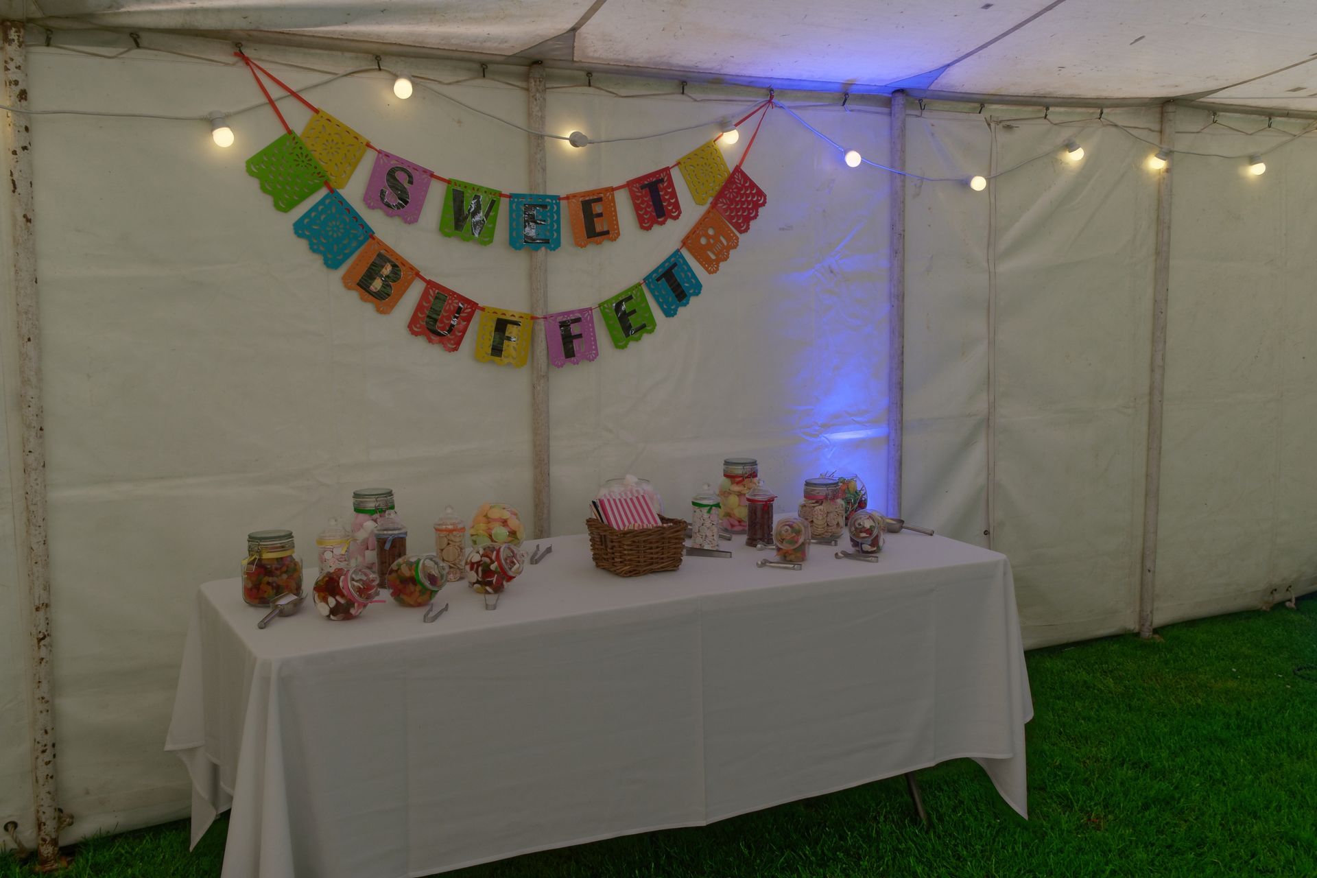 Sweet buffet table with jars of candy, a banner, and string lights inside a white tent.