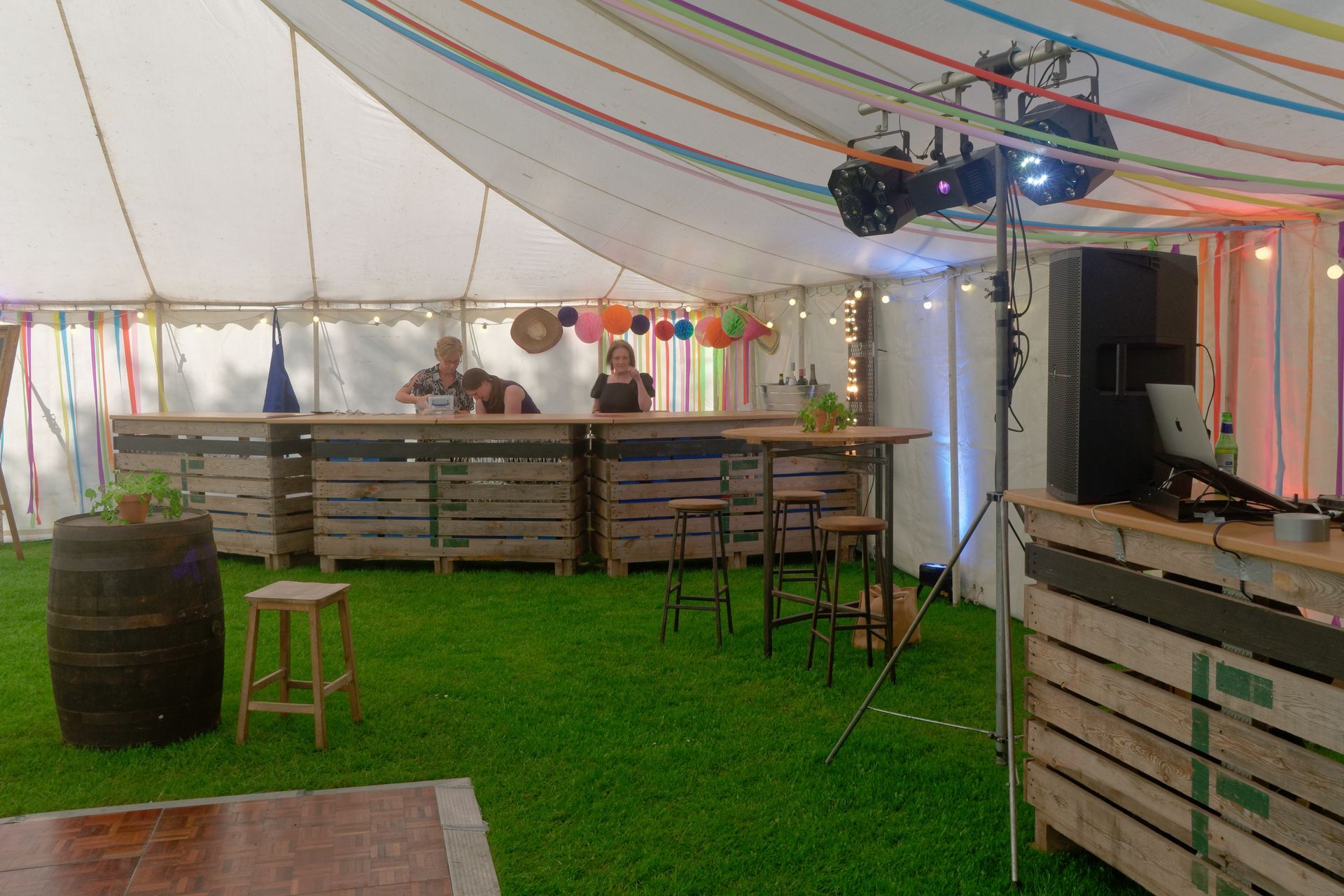 Bar setup inside a tent. People behind the bar, colorful streamers, artificial grass, and a wooden dance floor.