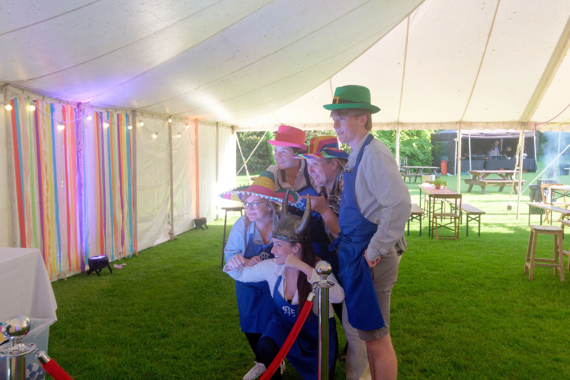People wearing hats pose for a photo in a photo booth, inside a marquee on a grassy lawn.