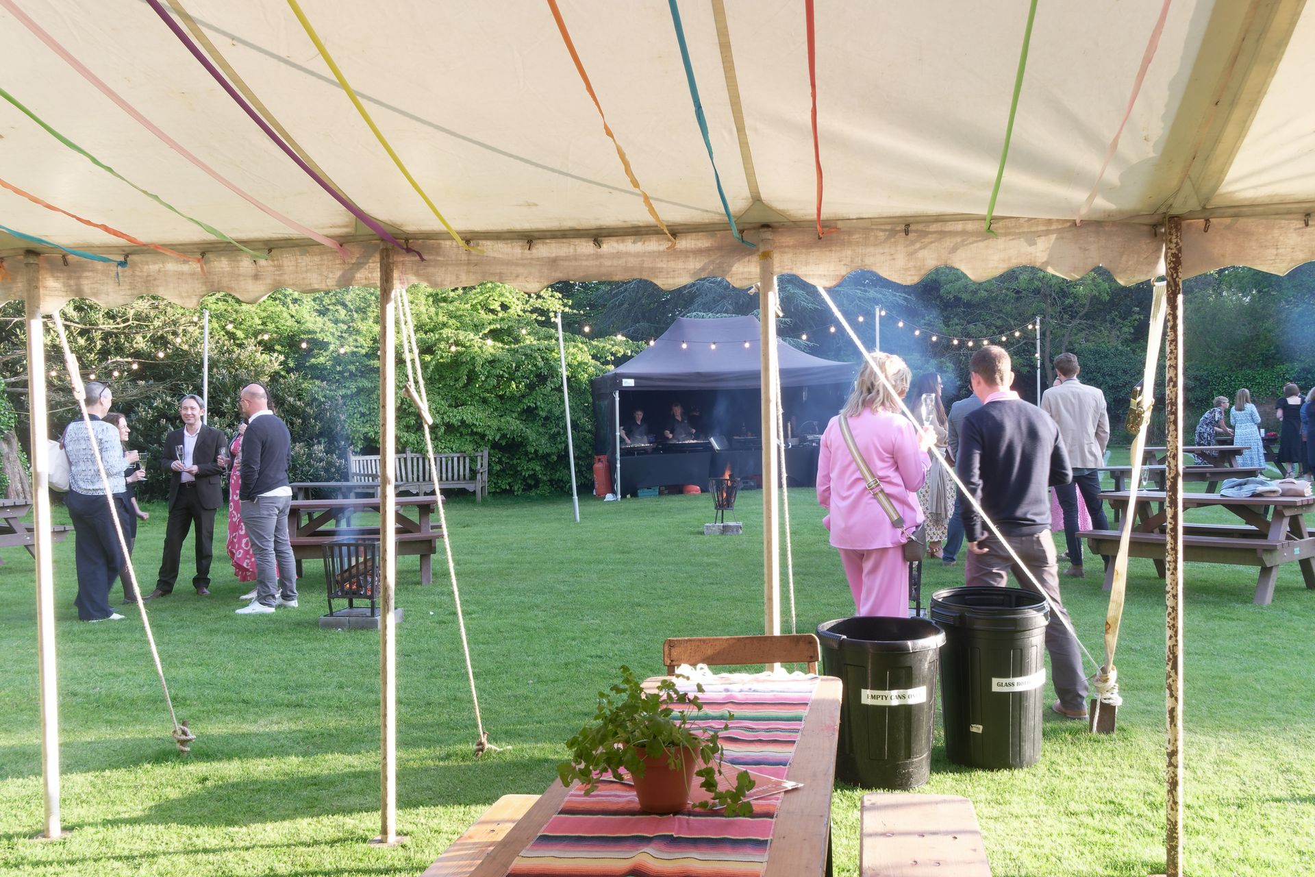 People socializing under a tent on a grassy lawn. Picnic tables, a black tent, and a fire pit are visible.