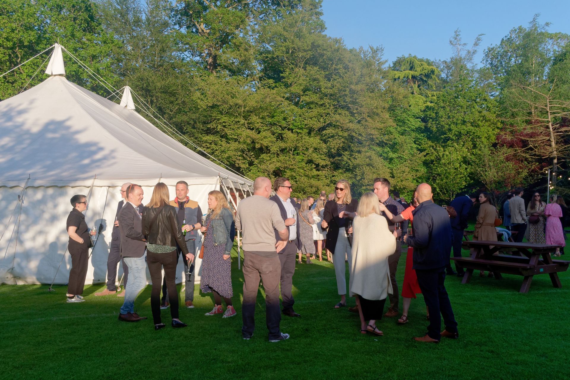 People socialize on a grassy lawn near a white tent. Sunlight filters through trees.