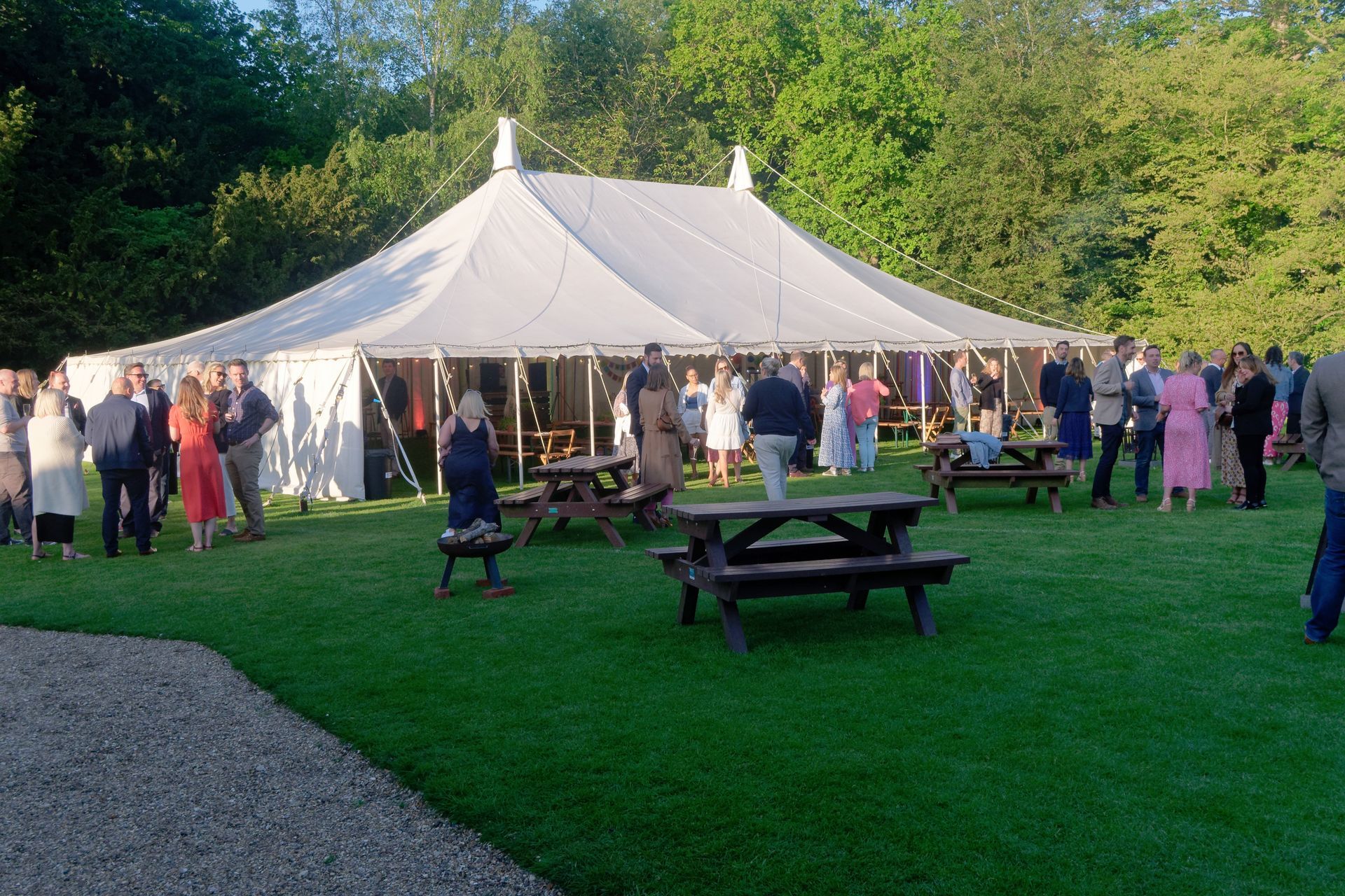 People gather outside a white tent on a grassy lawn. Picnic tables are in the foreground.