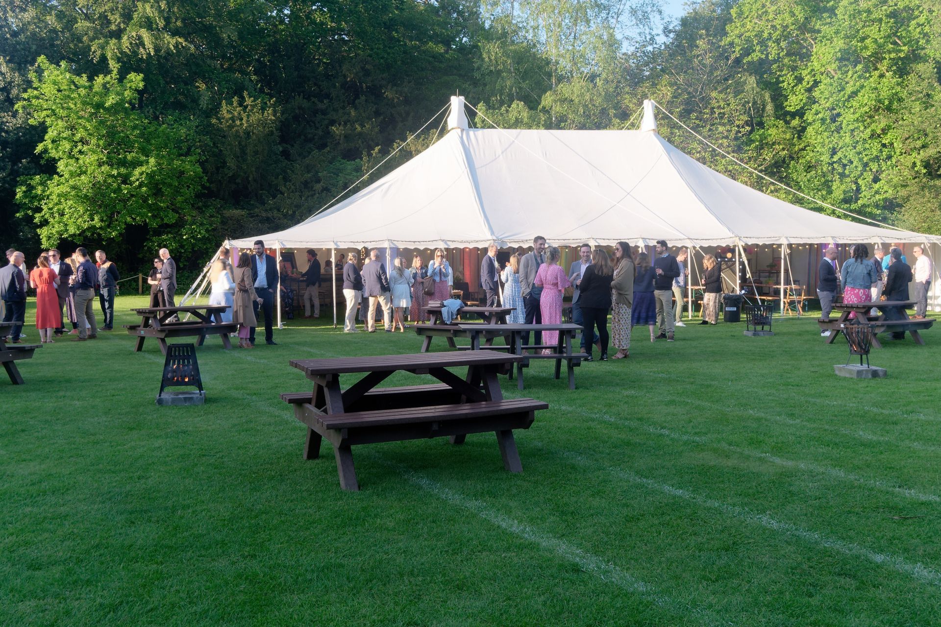 People gather outside a large white tent on a grassy lawn with picnic tables; trees in the background.