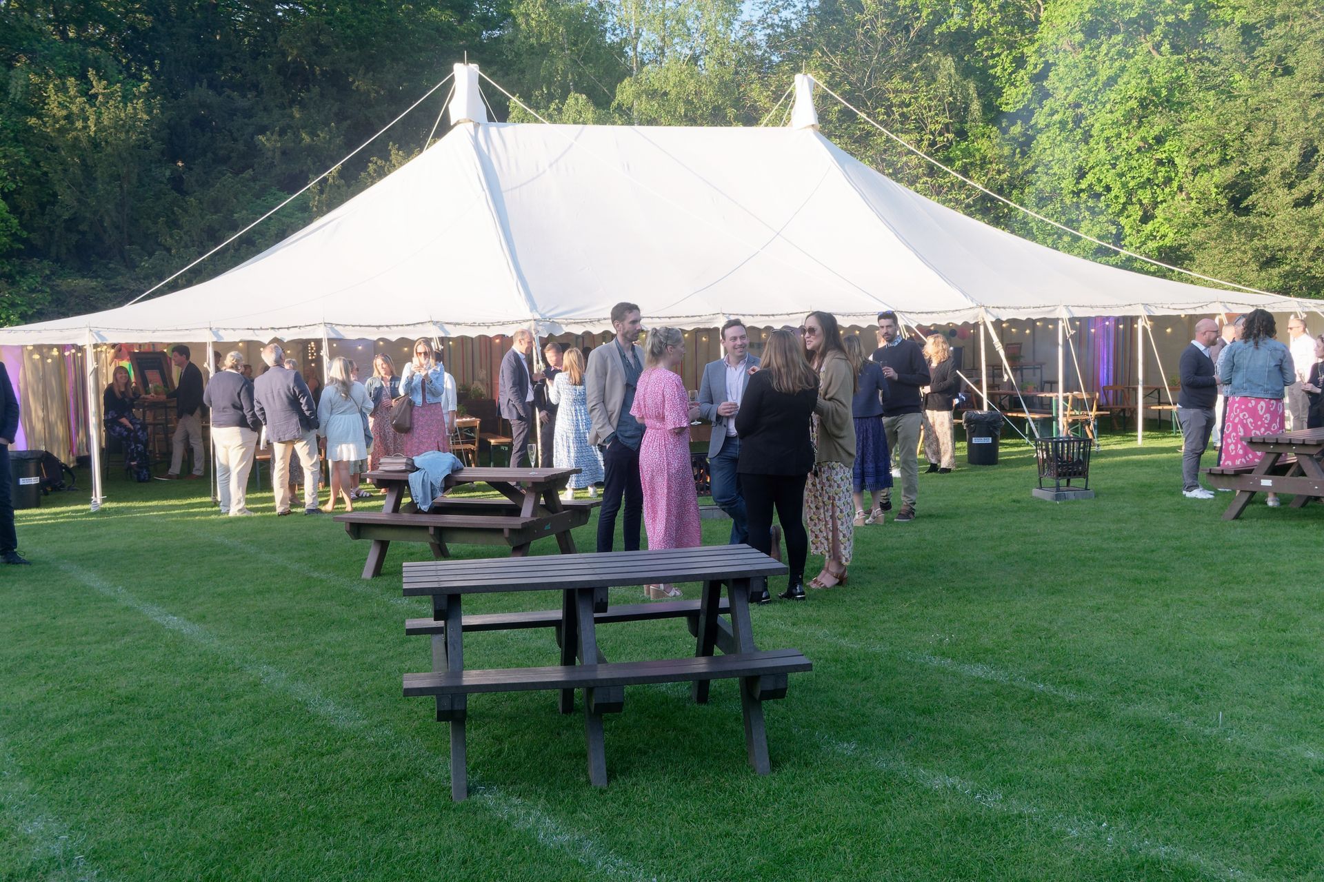 People mingling near a white tent in a grassy outdoor setting with picnic tables.