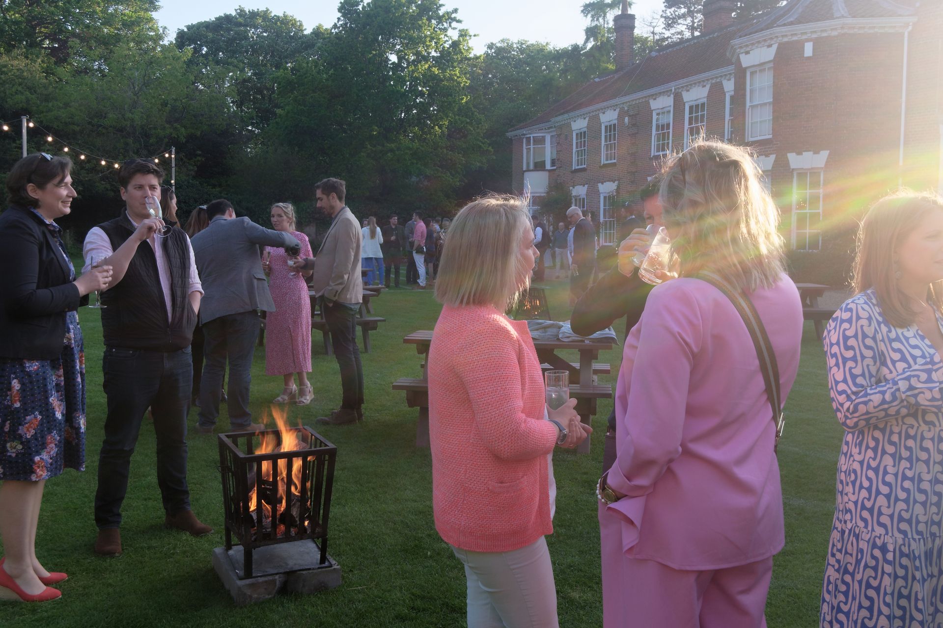 People gather outdoors at a party near a fire pit. Sunlit evening.