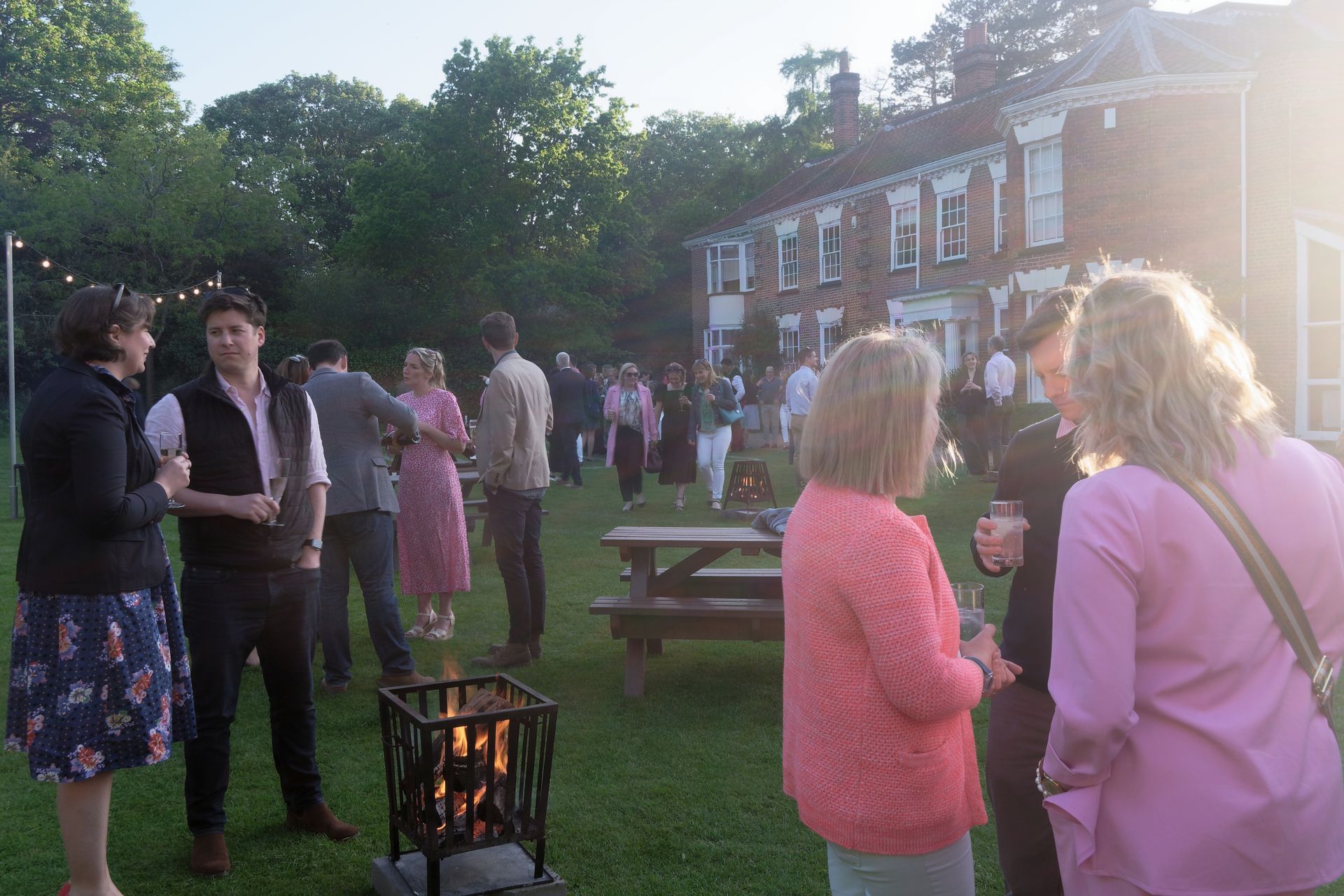 People socializing at an outdoor gathering on a grassy lawn, with a bonfire and a large house in the background.