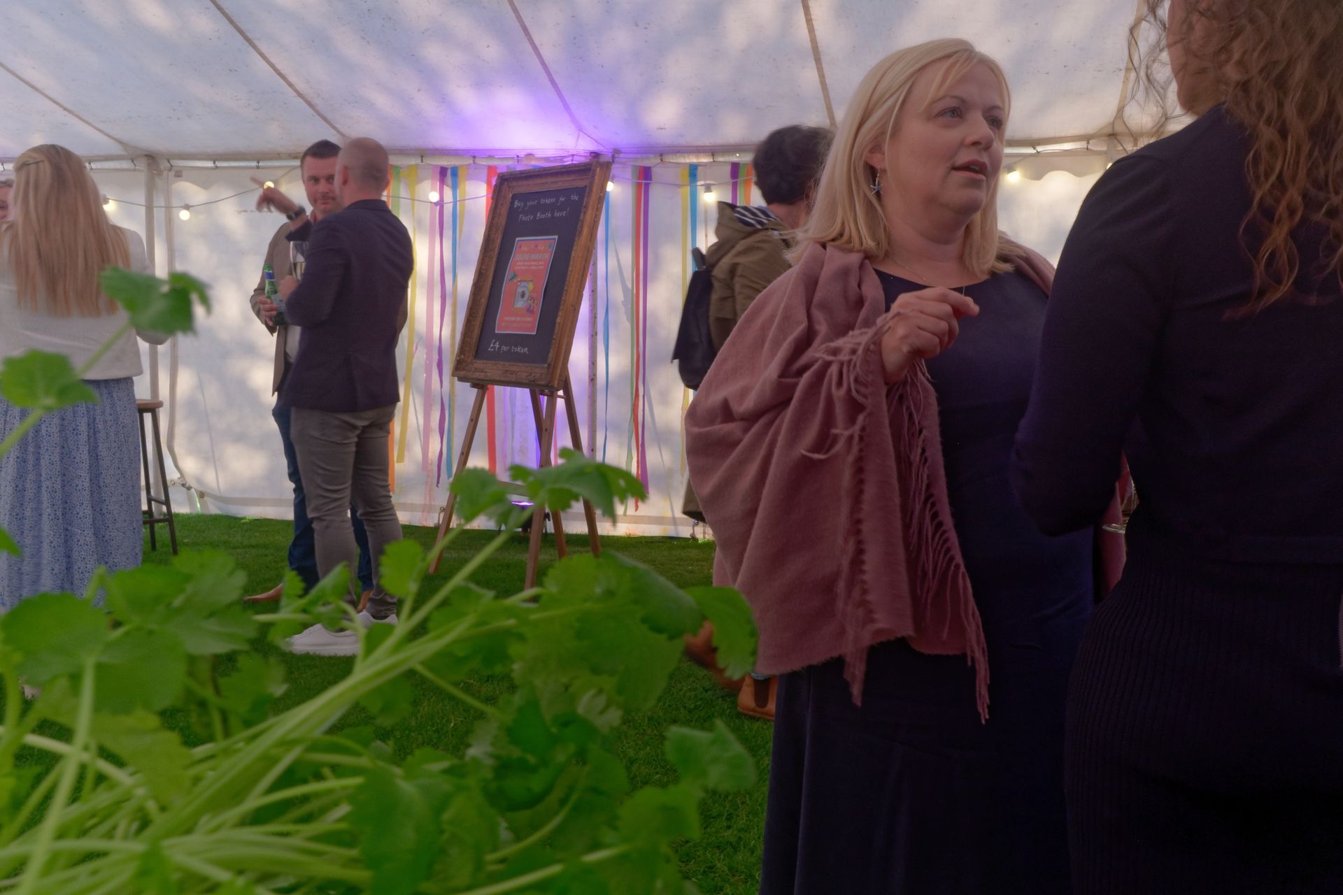 People inside a tent, woman in shawl conversing. A man plays a guitar, colorful streamers in background.