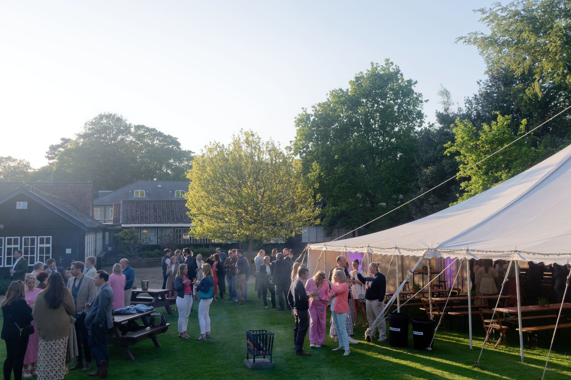 Large outdoor party with many people gathered near a tent, buildings, and picnic tables.