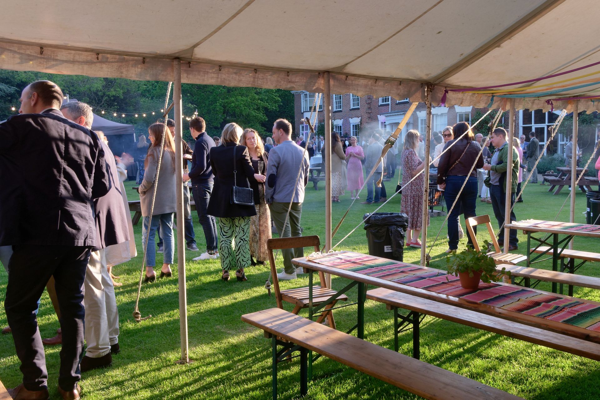 People gathered at an outdoor event under a tent with picnic tables and string lights.