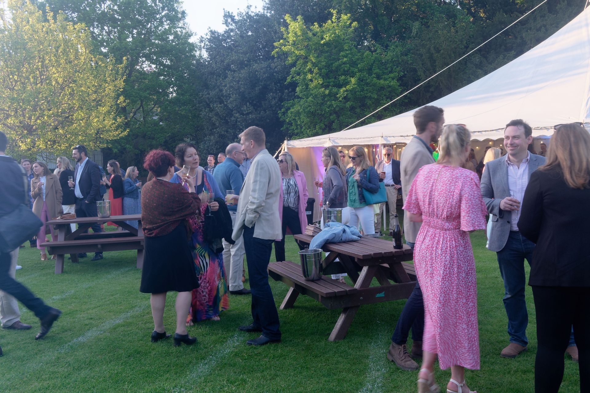 People socialize outdoors near a tent on a grassy lawn at dusk.