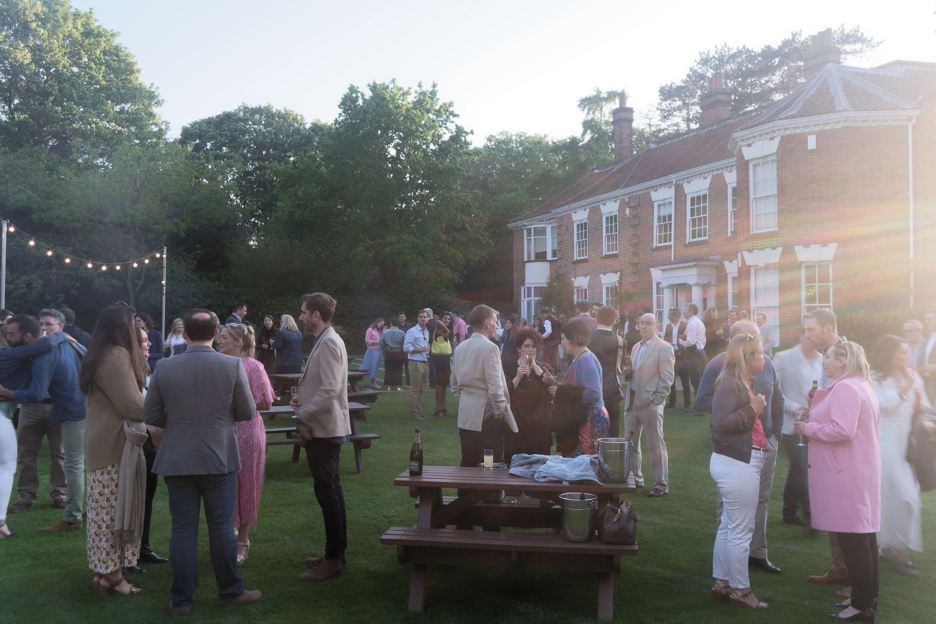 People at a party on a lawn in front of a brick building. Sunset with string lights.