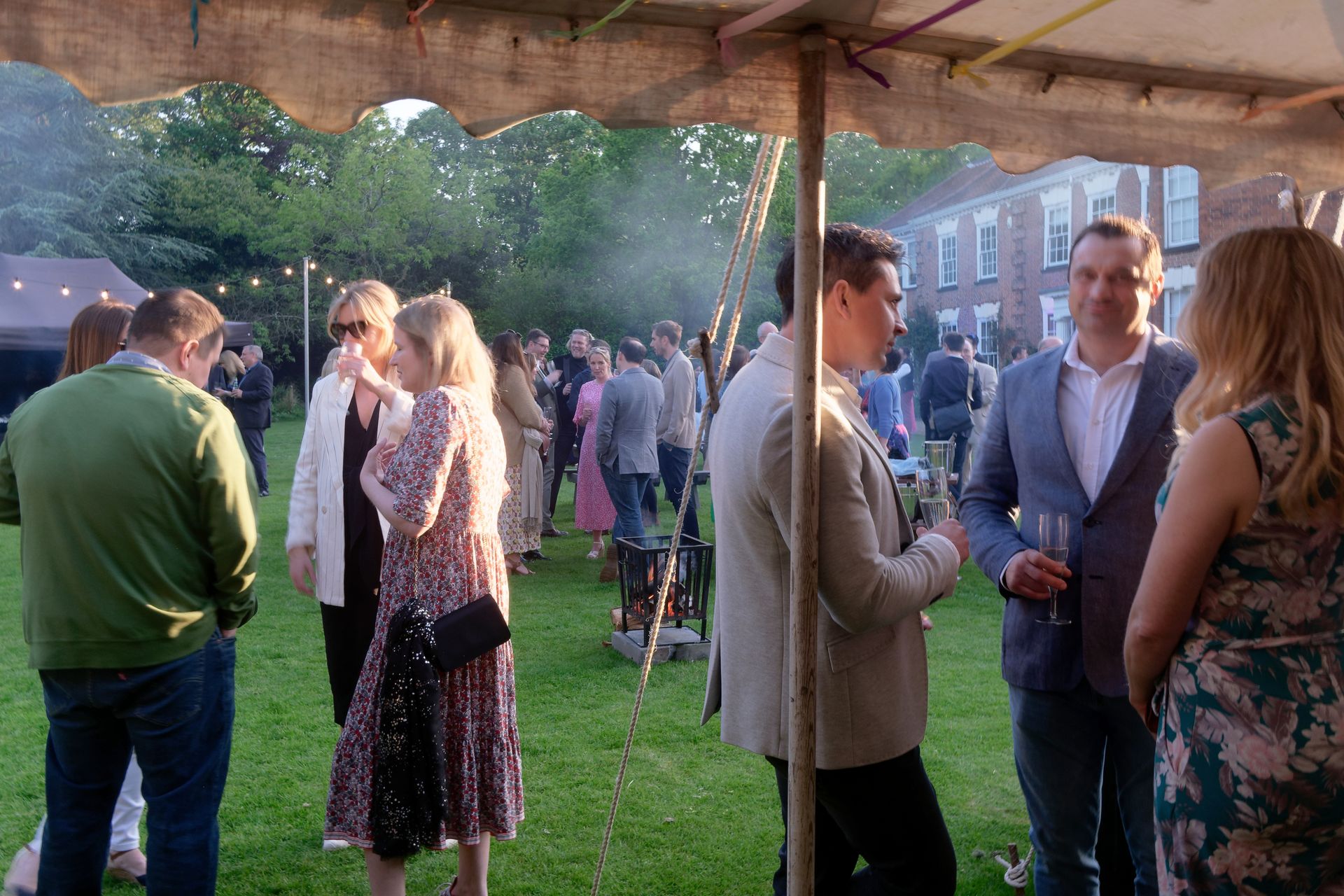 People socializing on a lawn under a tent. Evening event with drinks, greenery, and a brick building in background.