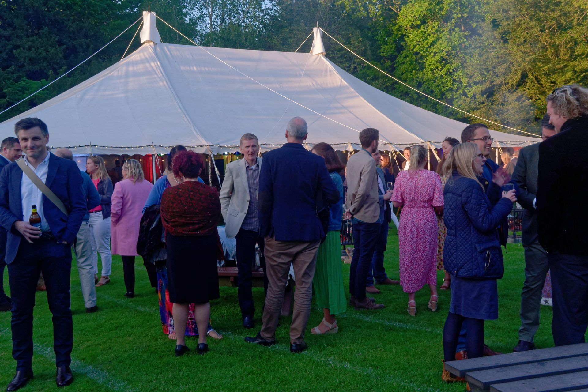 People mingling outdoors near a large white tent on a grassy lawn. Evening gathering.