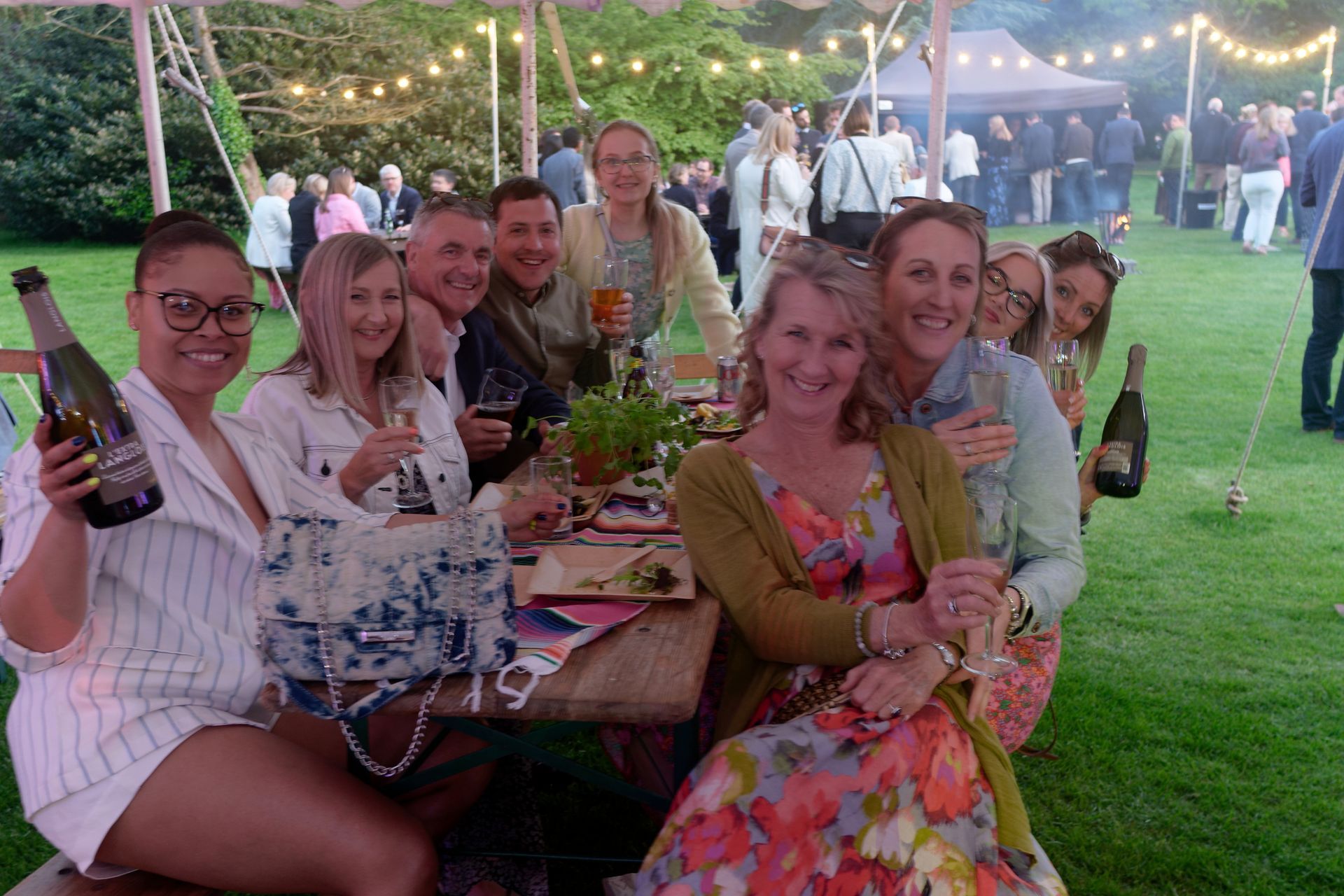 Group of people at an outdoor gathering, smiling at a table, holding drinks, and surrounded by green grass.