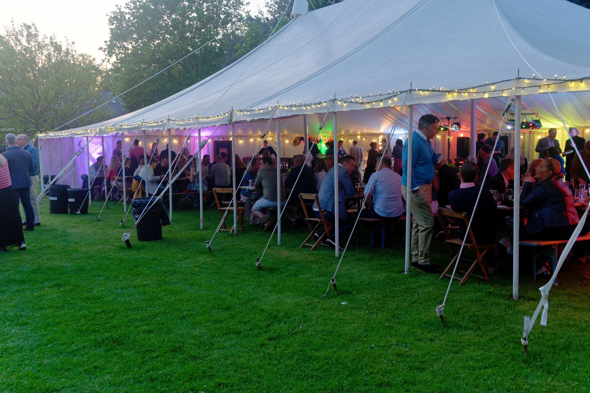 Large tent hosting an outdoor party with people seated at tables on a green lawn, illuminated by colorful lights.