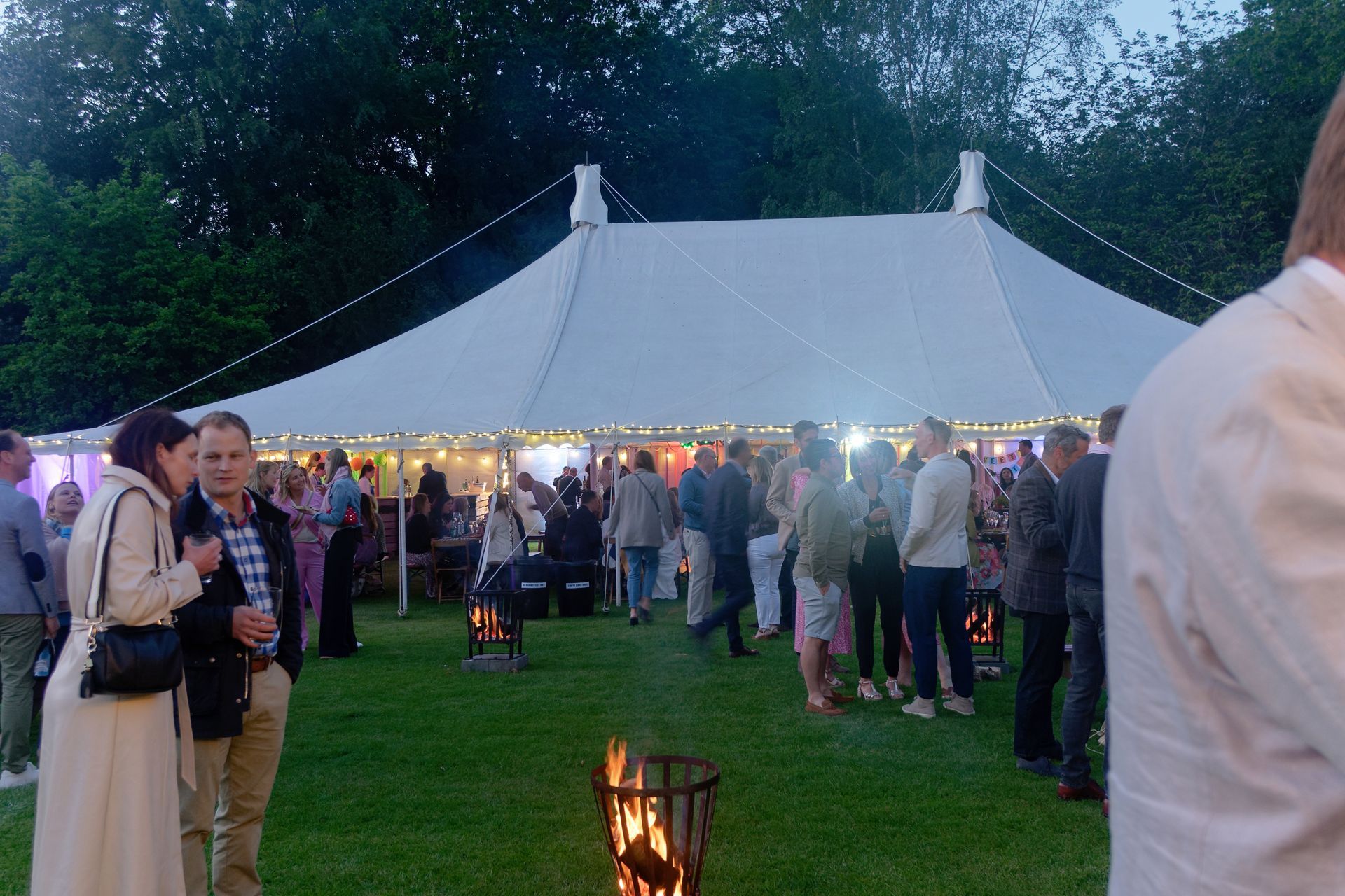 People at an outdoor party under a large white tent, with string lights, on a grassy lawn.