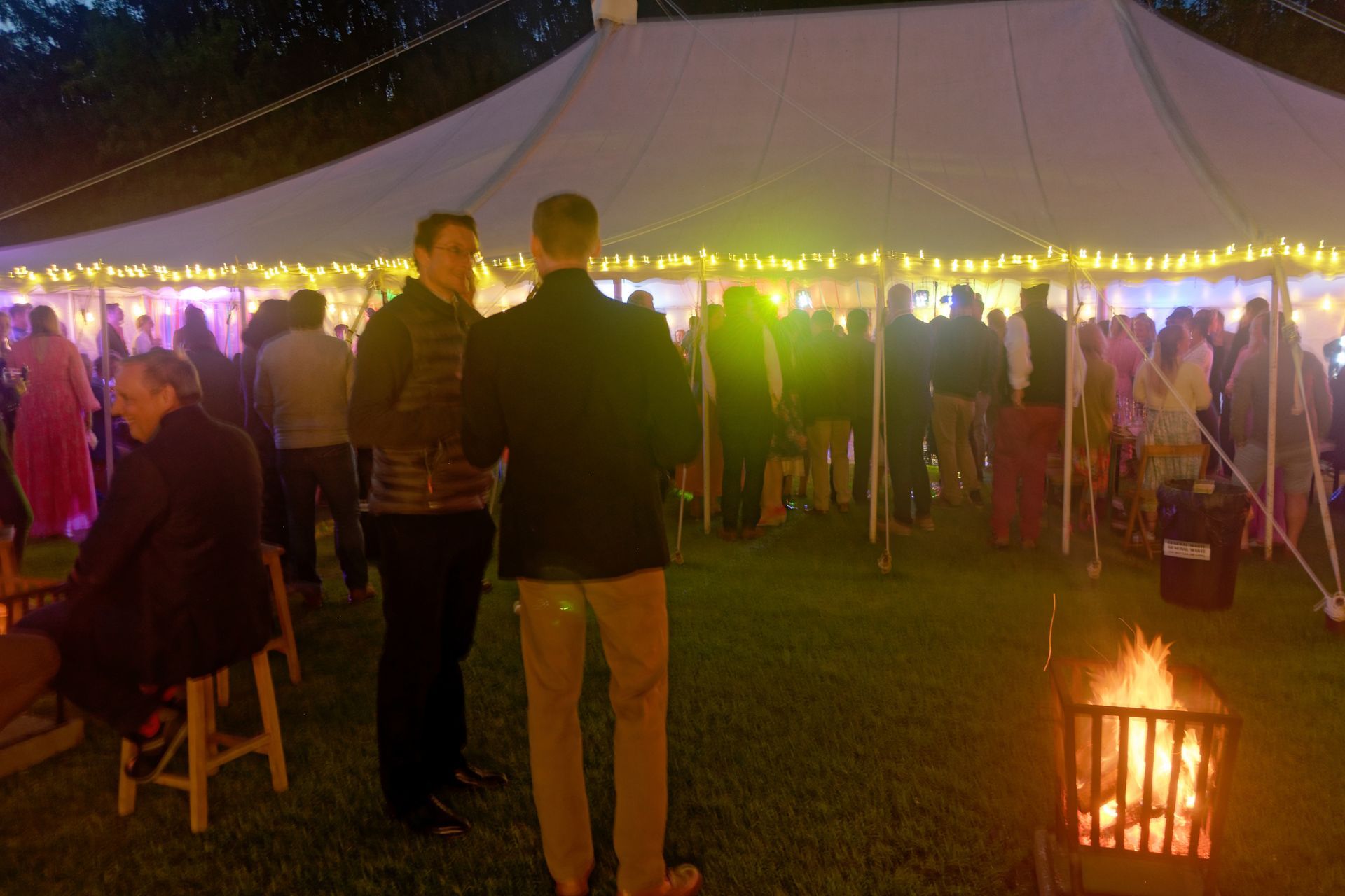 People at a party inside and outside a white tent, lit with string lights. A bonfire is in the foreground.