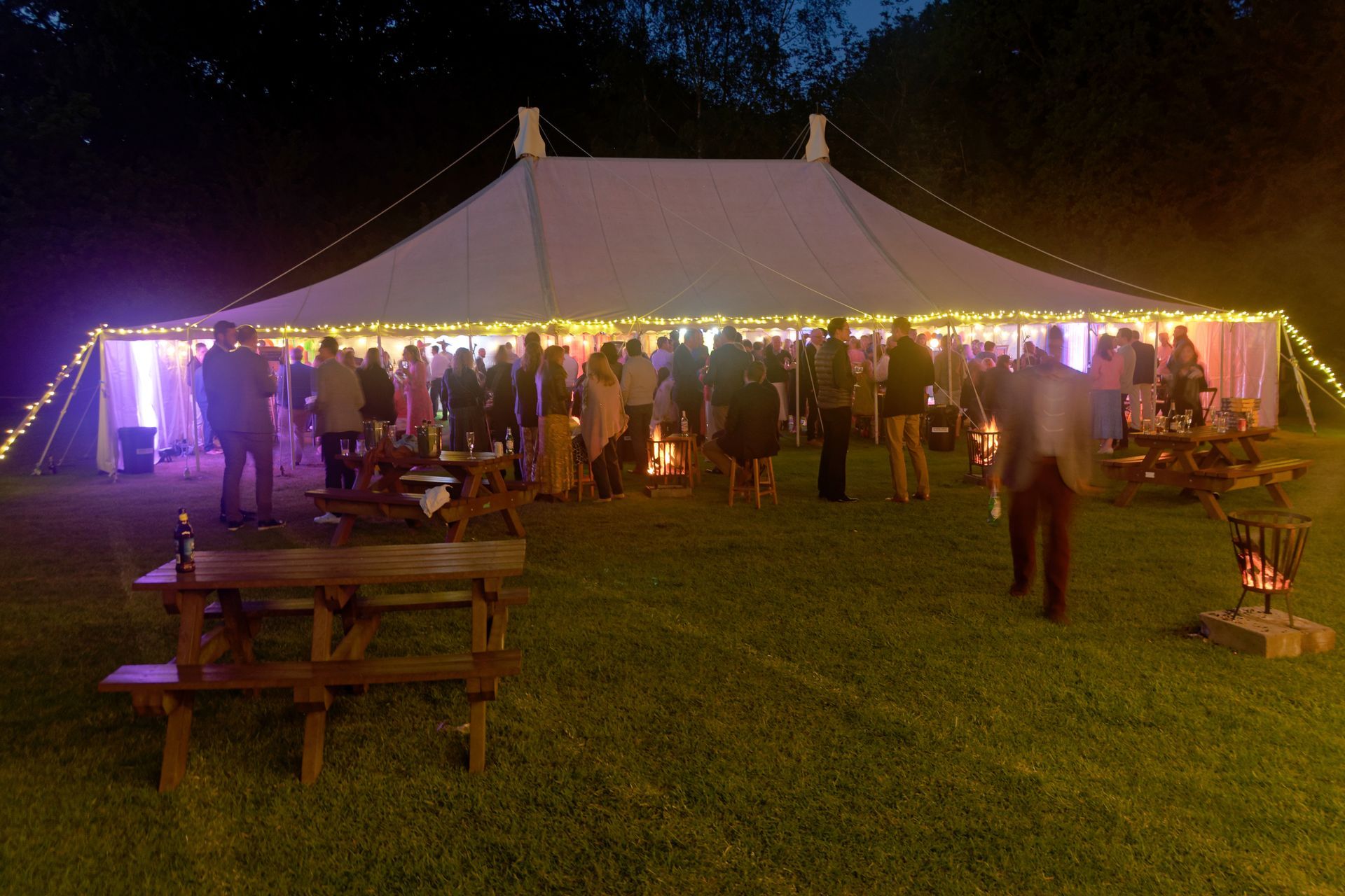 Outdoor party at night under a lit white tent with guests, picnic tables, and fire pits on the grass.