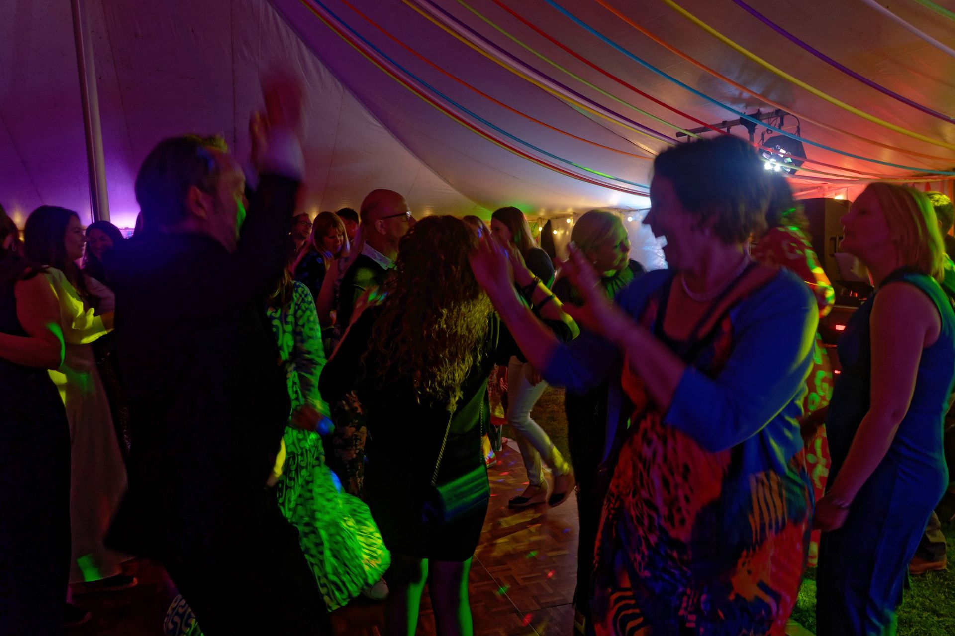 People dancing at a party inside a tent, colorful lights illuminating the crowd.