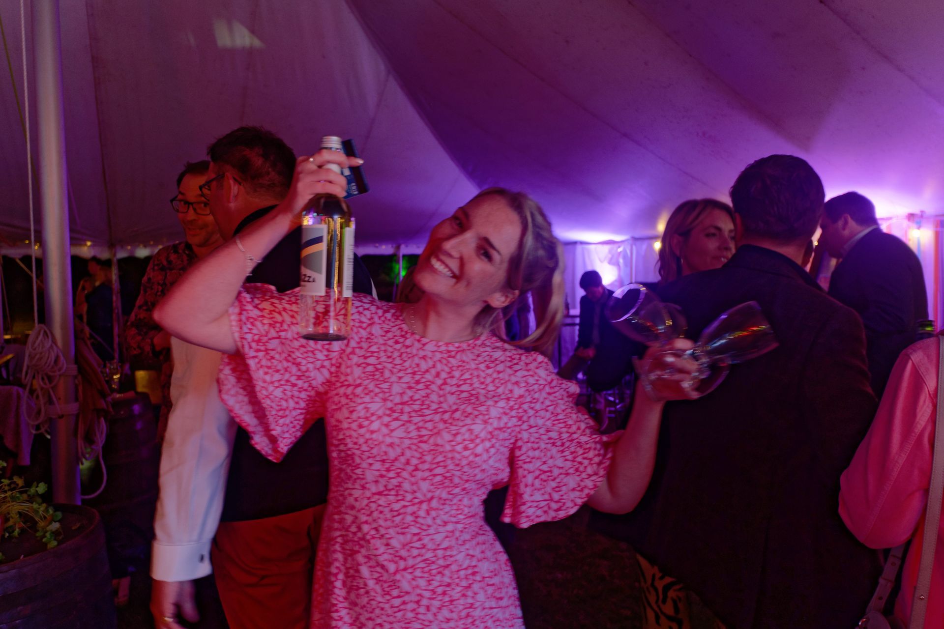 Woman in pink dress smiling and holding a bottle and wine glasses at a party.