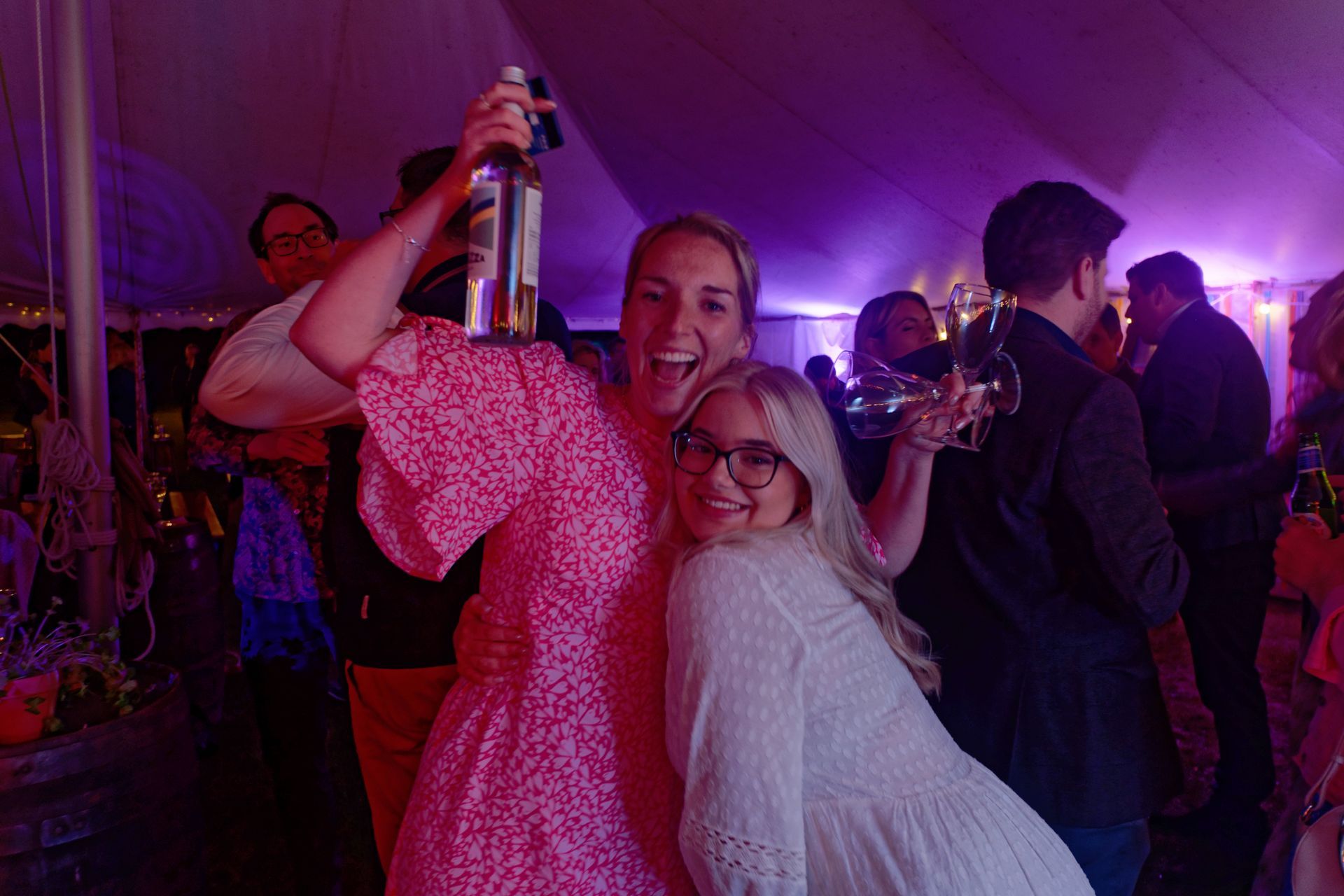 Two women laughing, one holding a bottle in a tent, surrounded by people at a party.