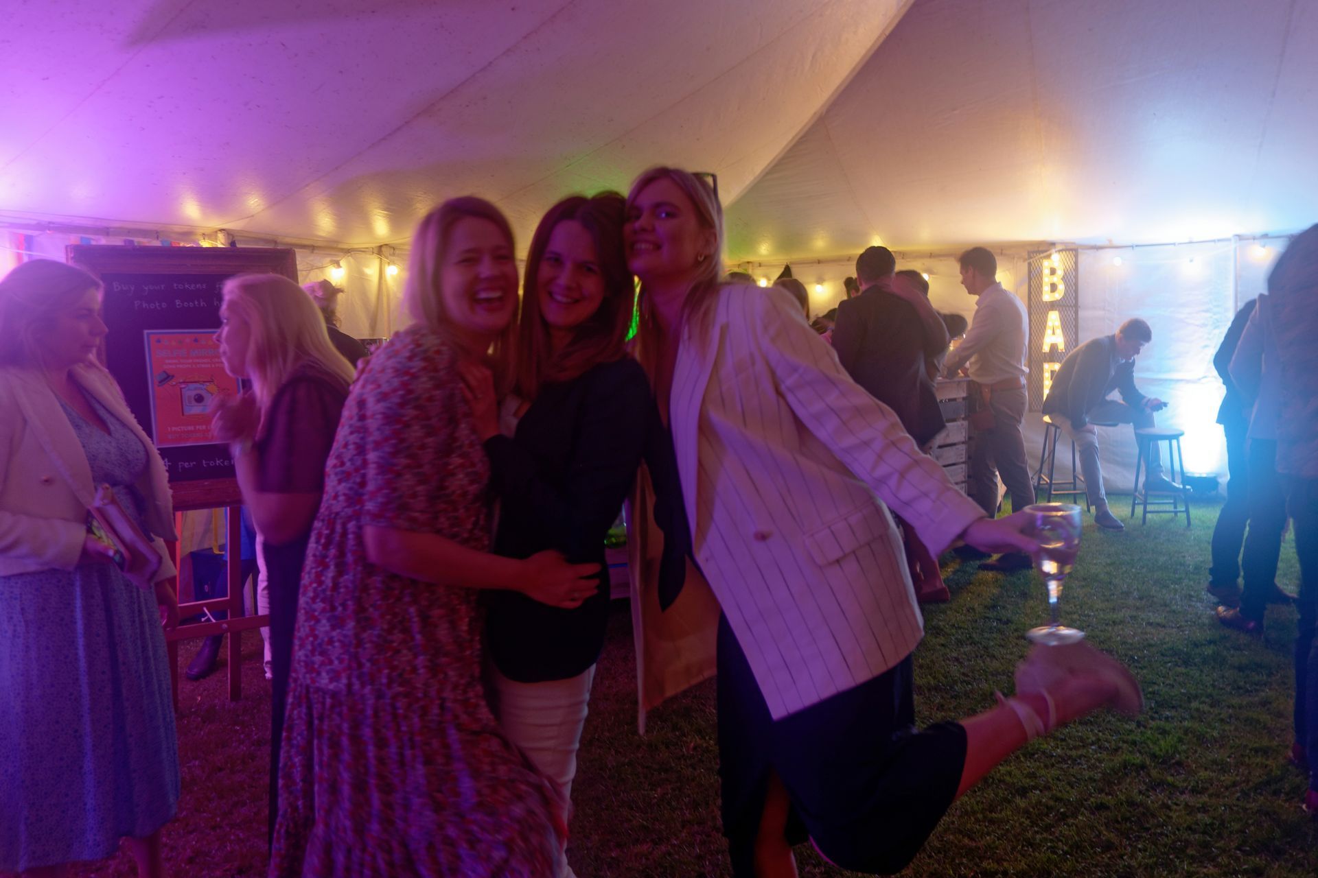 Three women posing, smiling, at a party under a tent.  One lifts her leg. People in background.
