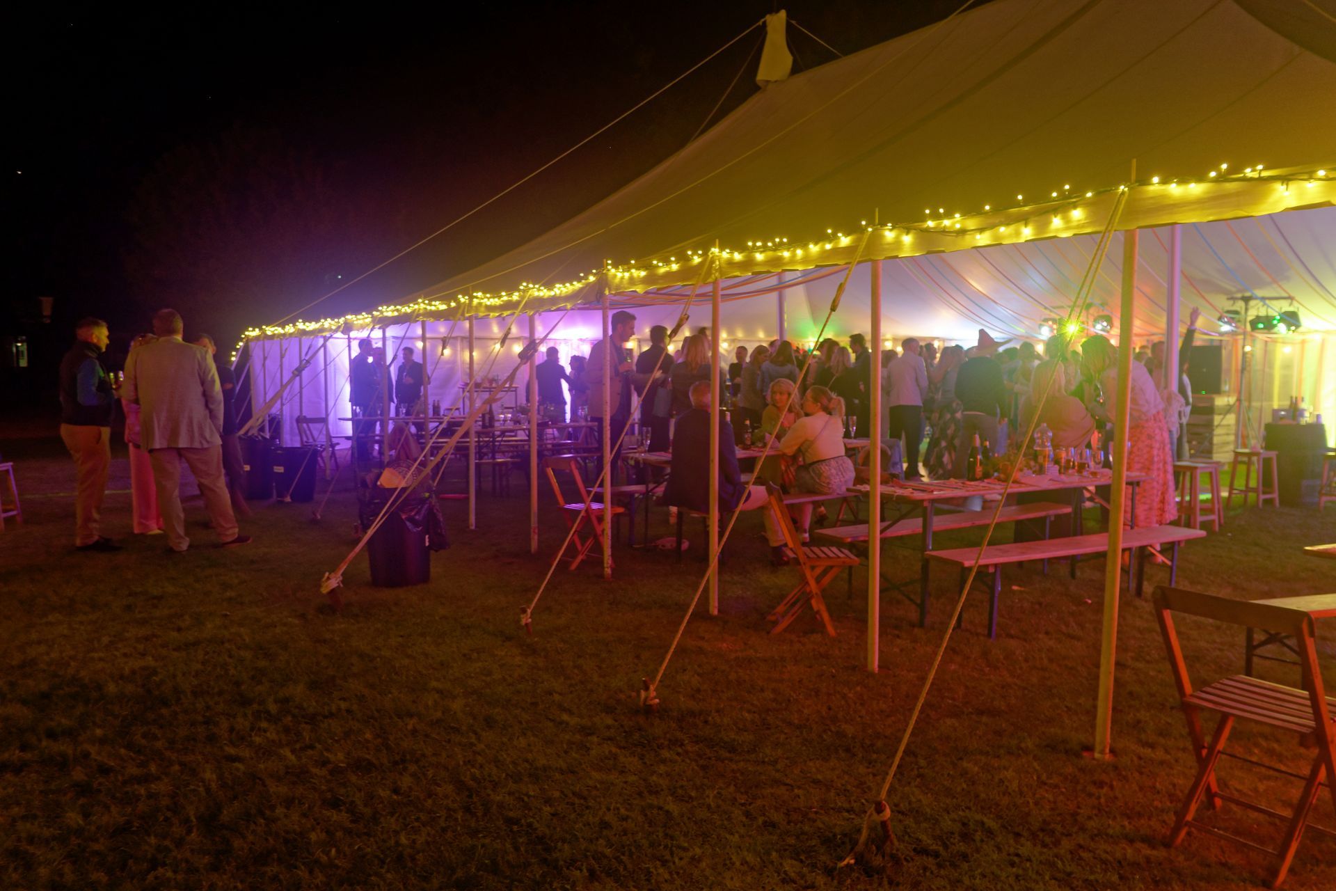 Nighttime outdoor party under a tent with string lights. People socialize around tables.