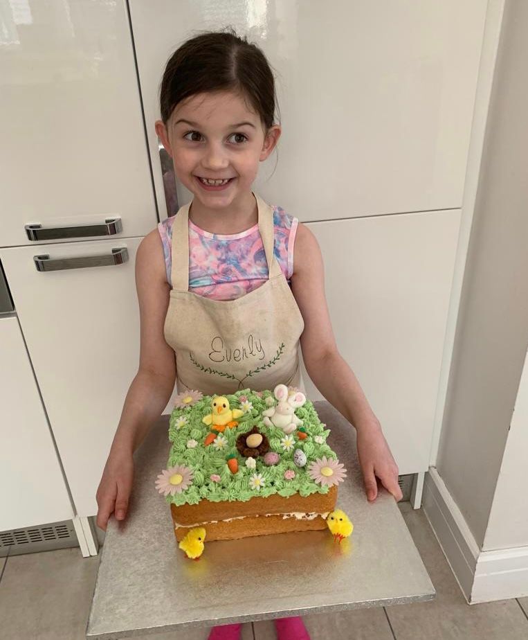 Young girl smiling, holding Easter cake decorated with chicks, grass, a nest and a rabbit.