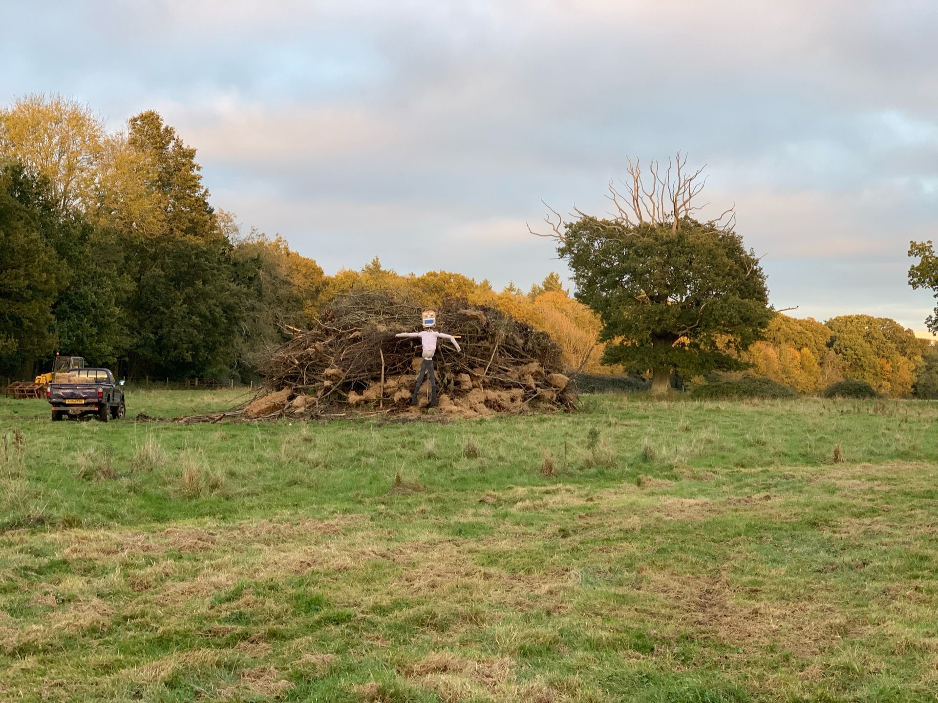 A scarecrow atop a large brush pile in a grassy field; a small tractor in the background.