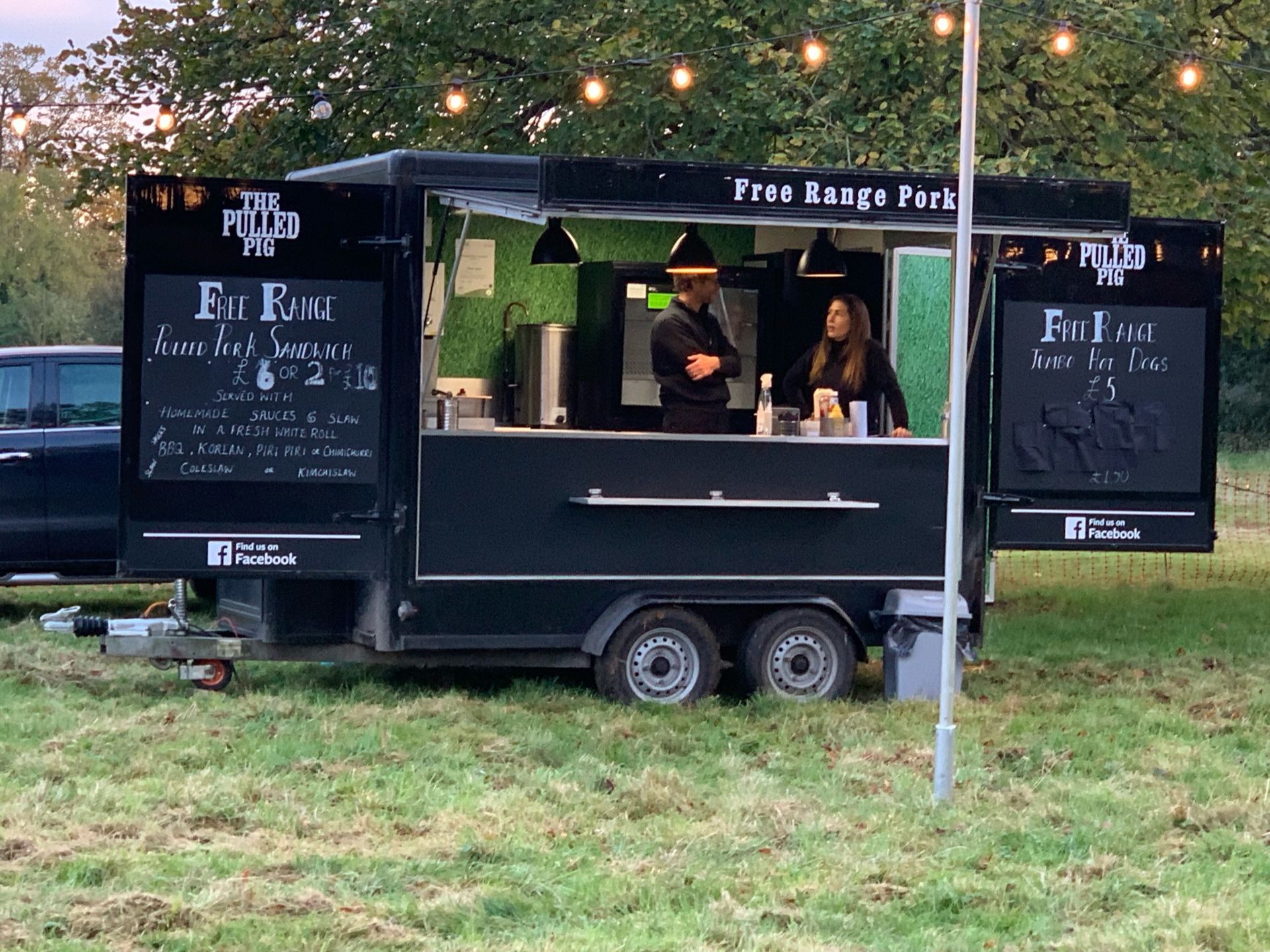 Black food truck in field, two people behind the counter. String lights overhead, green grass.