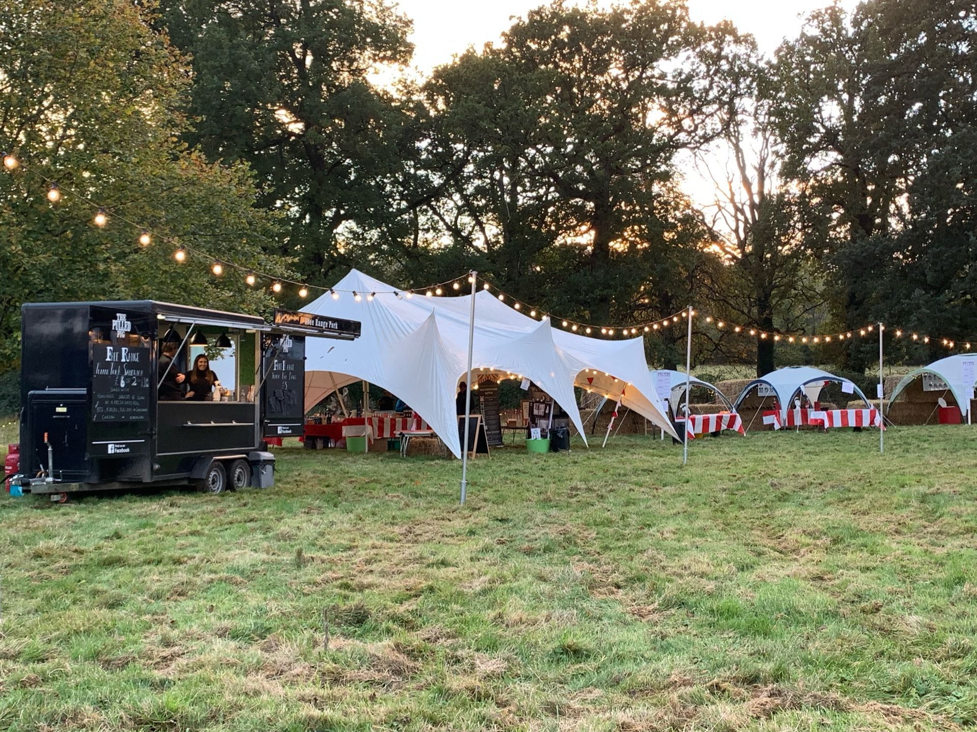 A food truck, tents, and string lights set up on a grassy field at dusk.