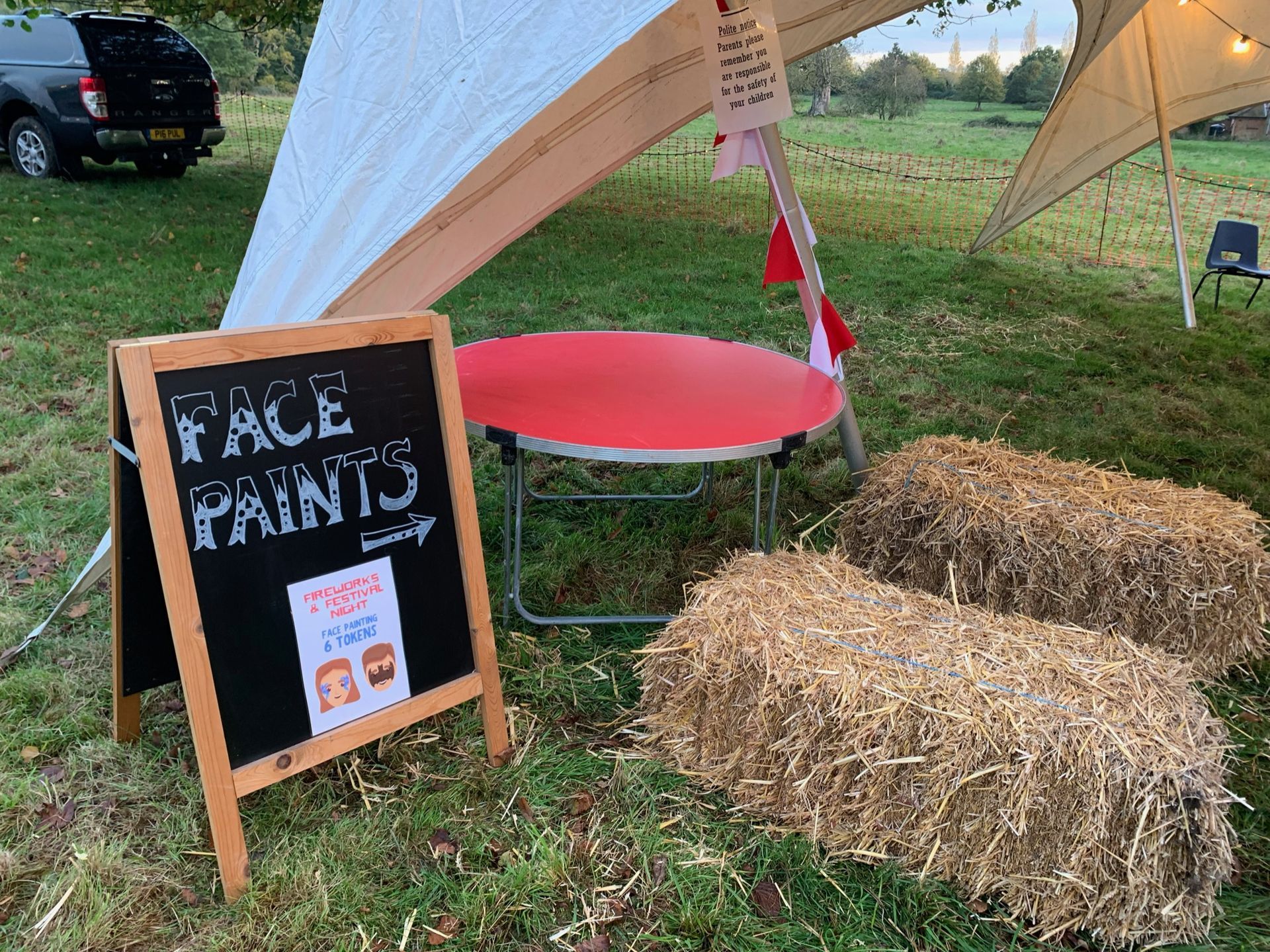 Face paint station: A-frame sign, red stool, hay bales, and a tent on a grassy field.