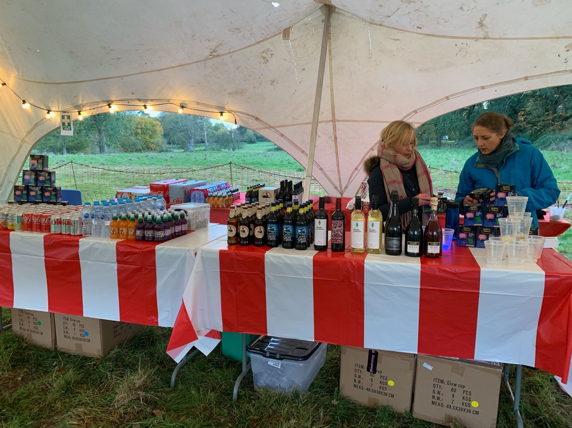 Two women serving drinks at an outdoor stall under a white tent with red and white striped tablecloth.